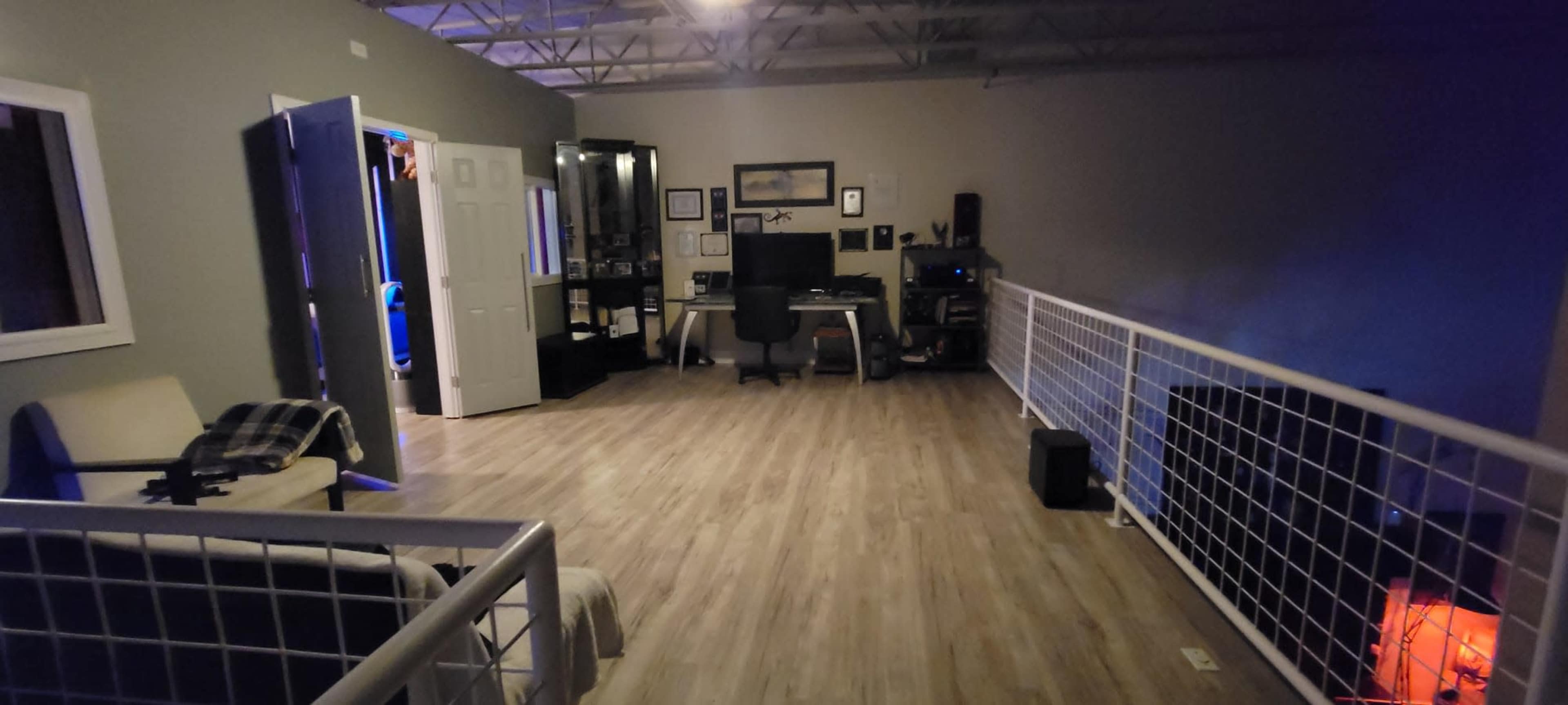 The image shows a spacious room with a desk and chair on one side, a couch with a blanket, and an open doorway leading to another area.