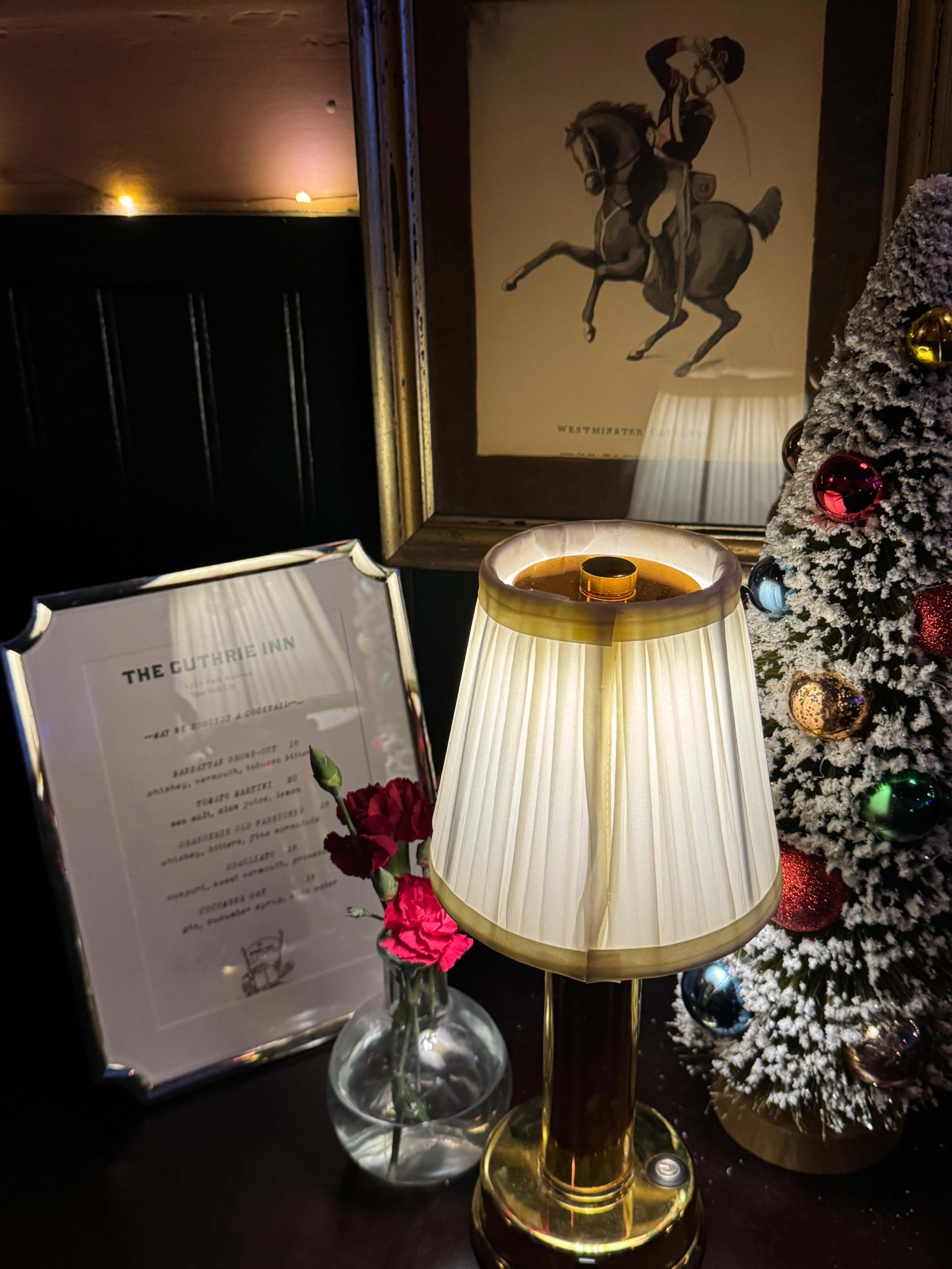 The image features a warmly lit table with a lamp, a vase of red roses, a framed menu or sign, and a small decorated Christmas tree.