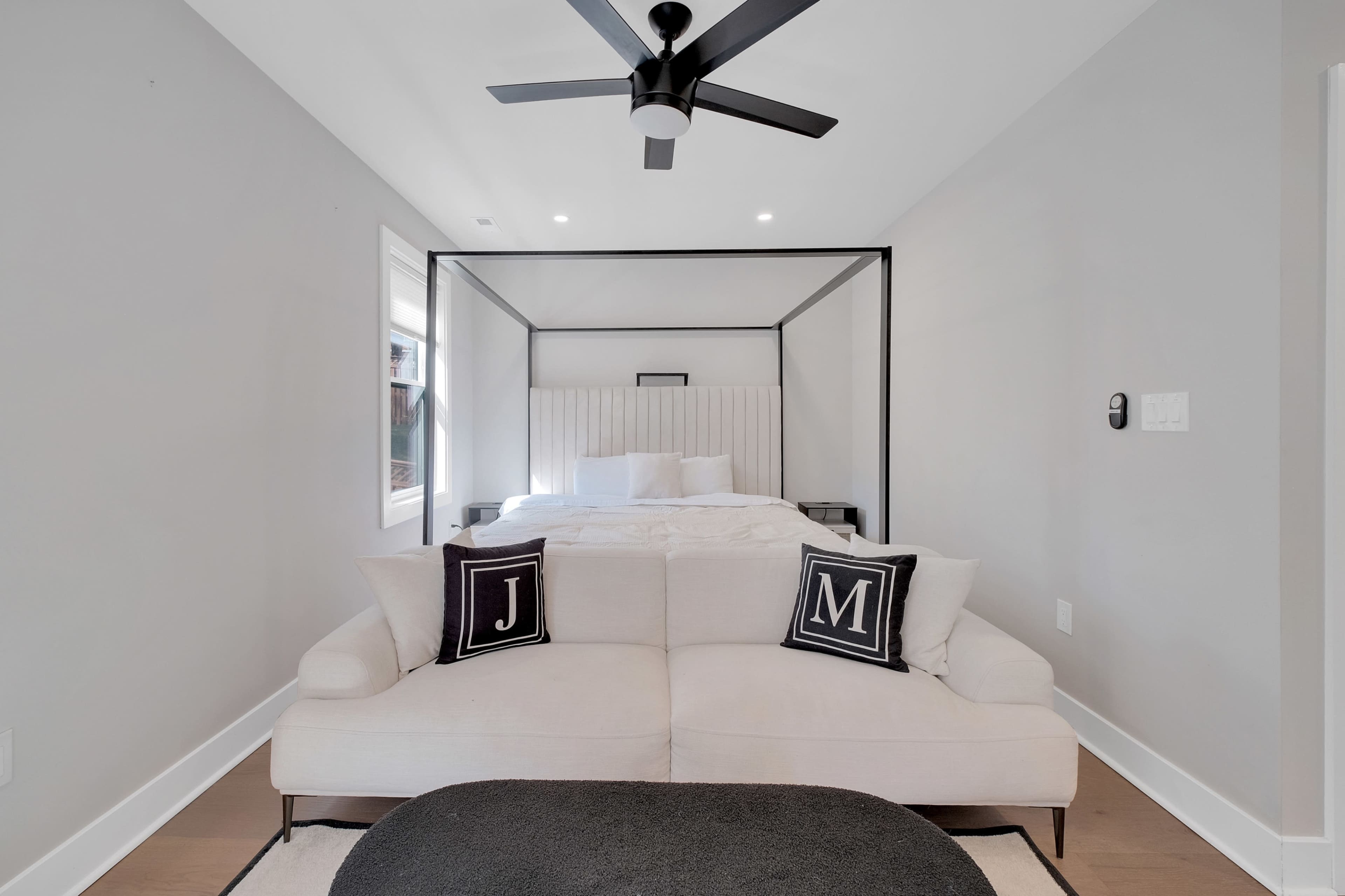 The image shows a modern bedroom featuring a four-poster bed, a light-colored sofa with decorative pillows, and a ceiling fan.