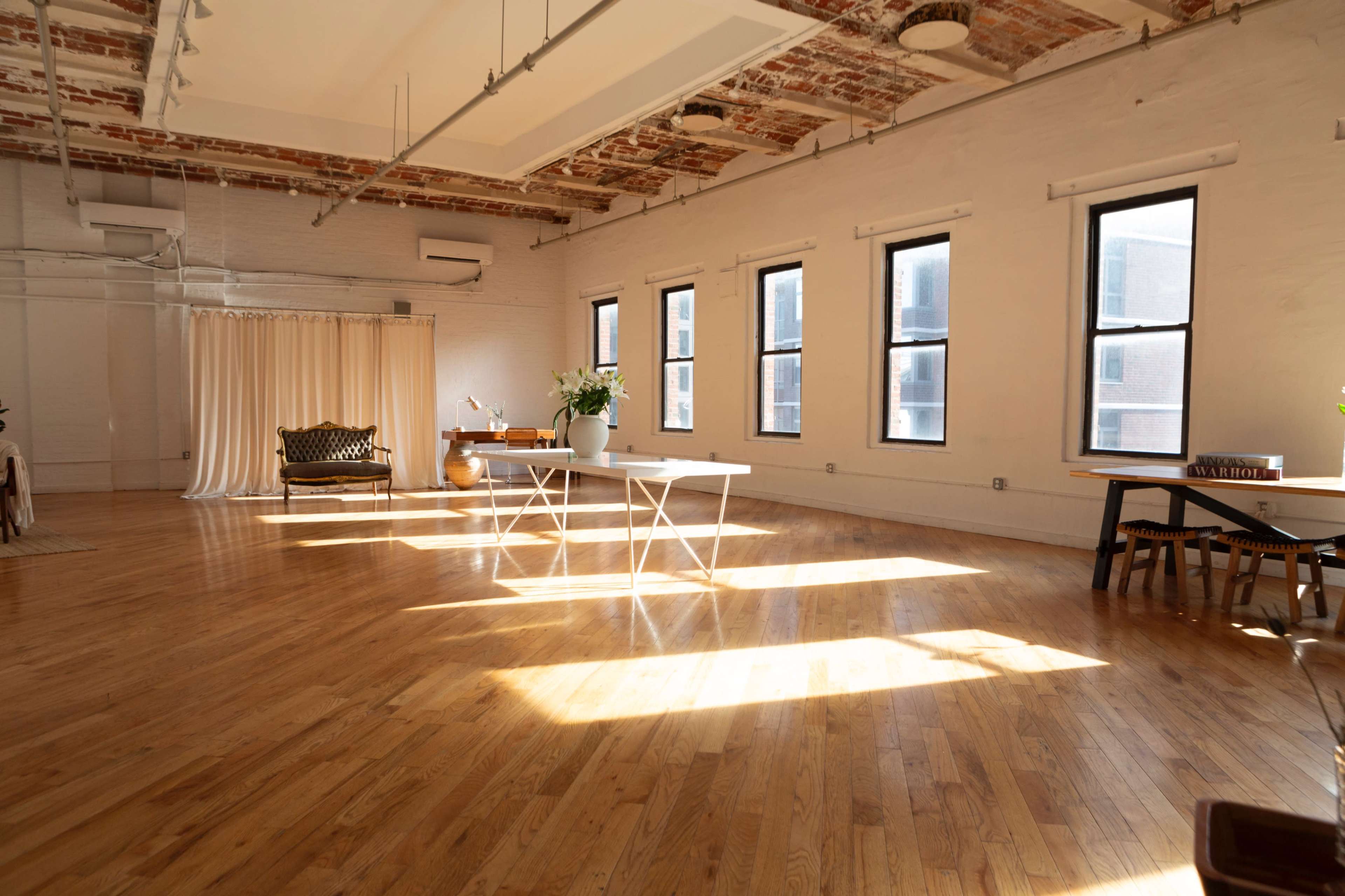 The image shows a spacious, well-lit room with wooden flooring, featuring large windows, a table, and minimal furnishings.