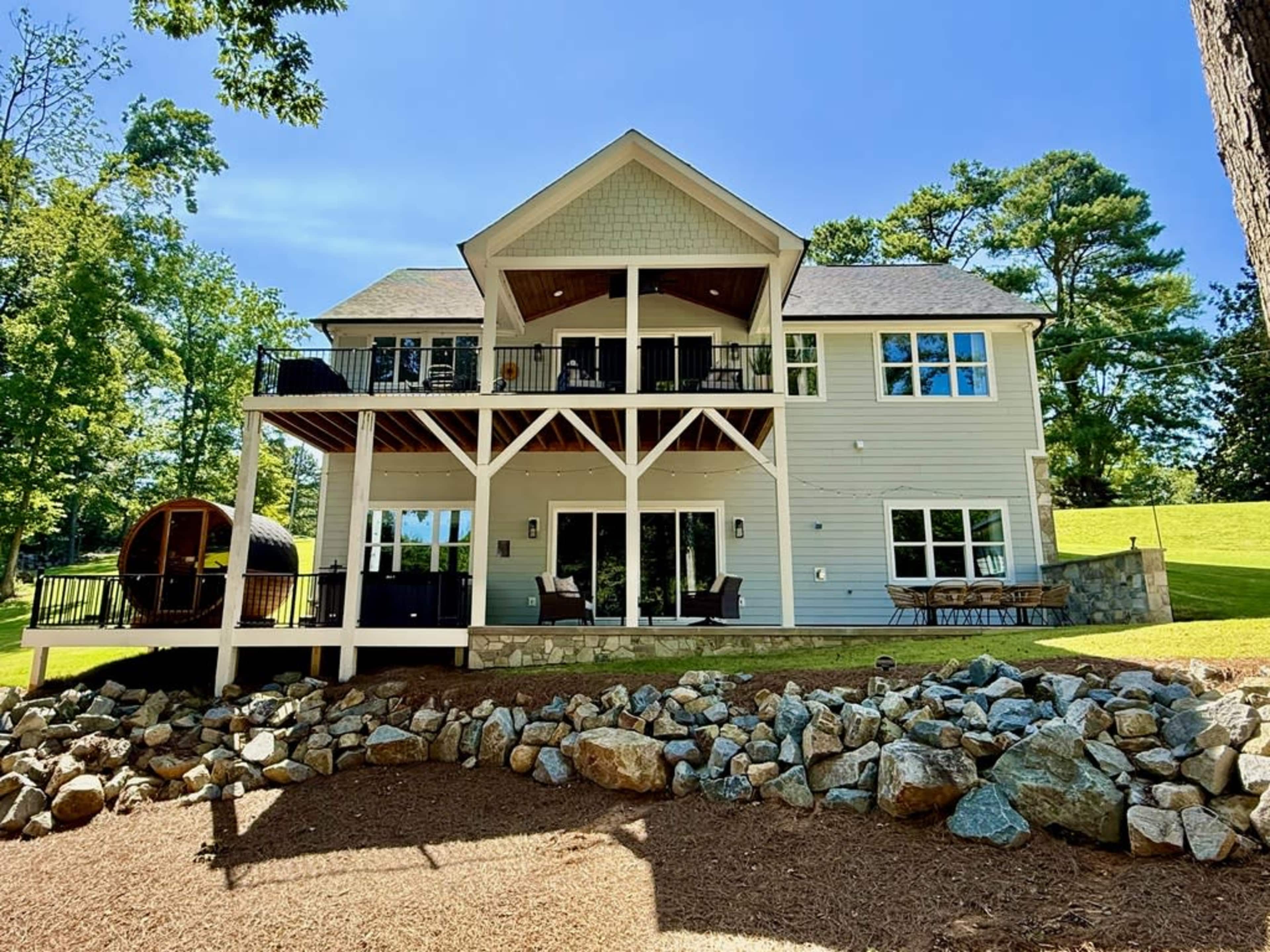 A two-story house with a wooden deck and large windows overlooks a landscaped yard with rocks and trees.