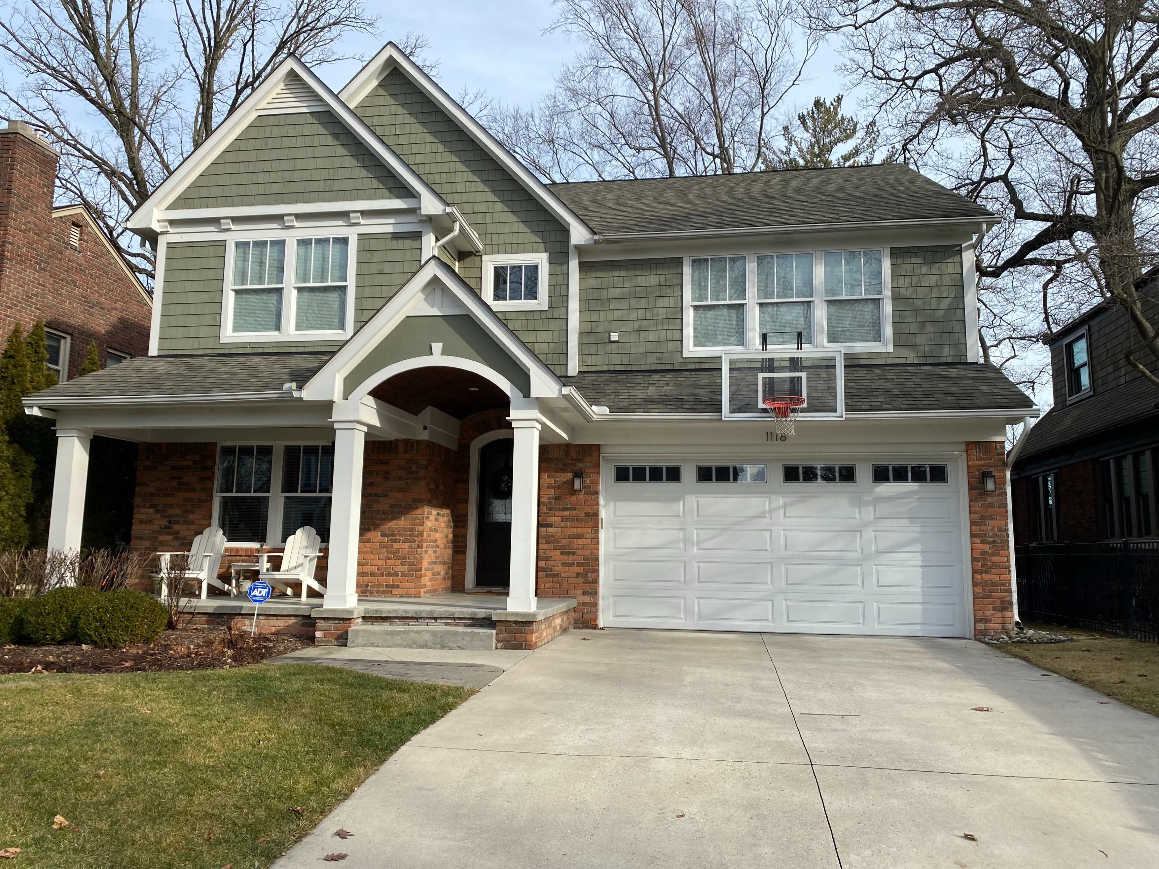 A two-story house with gray siding and brick accents features a front porch and a basketball hoop mounted on the garage.