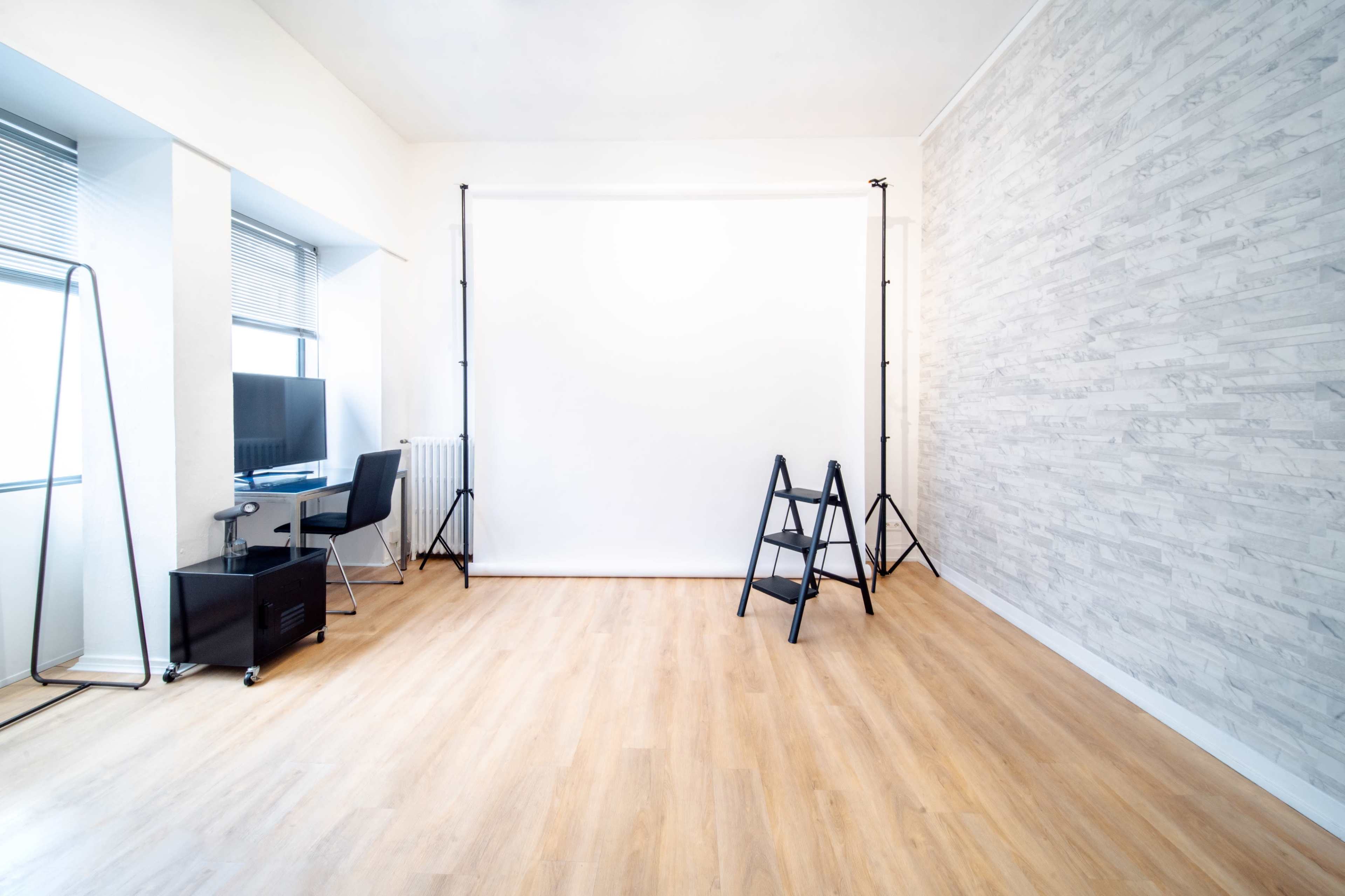 A bright, empty studio features a white backdrop on stands, a chair, a small desk with a monitor, and a ladder against a light-colored wall.