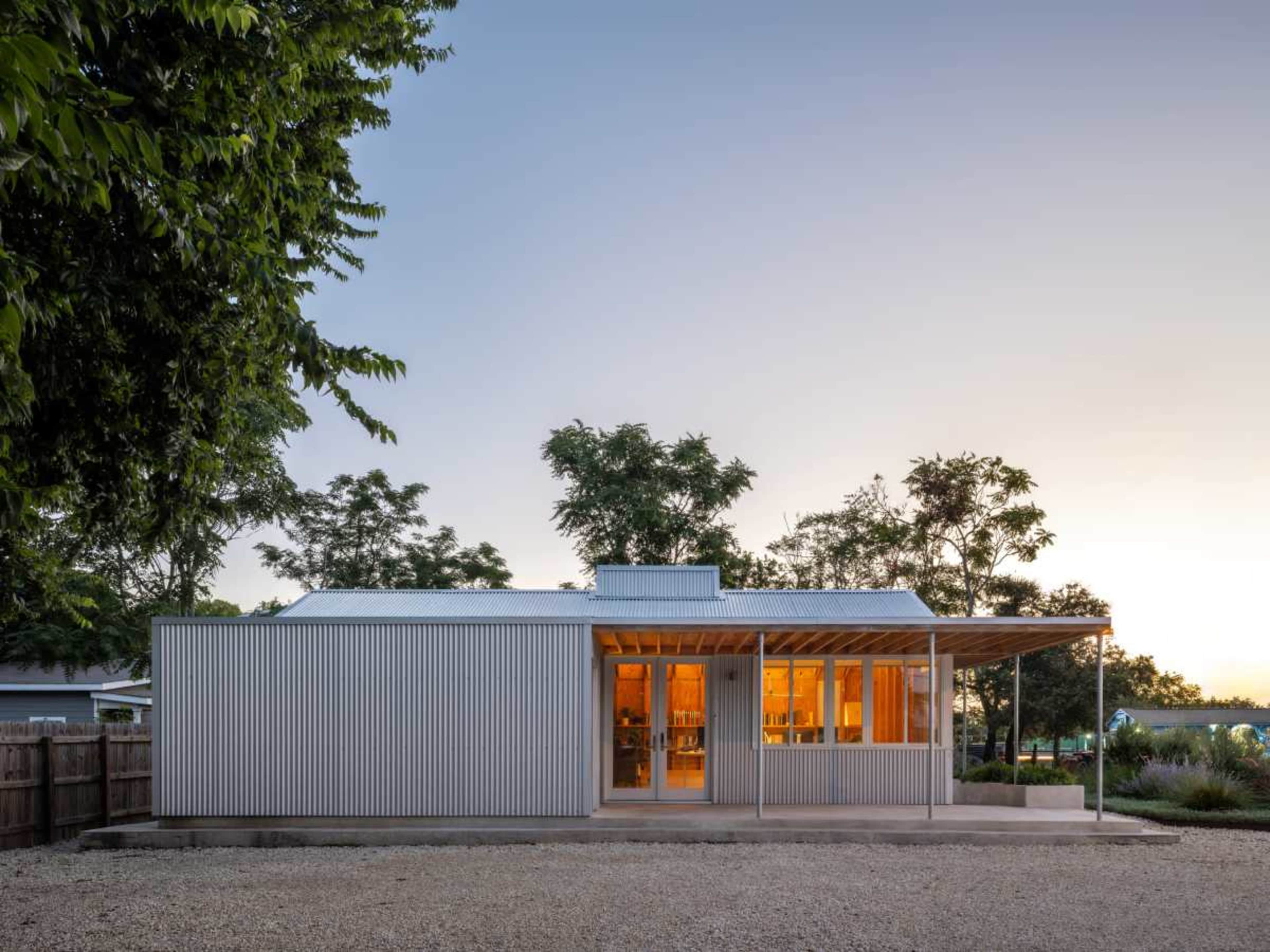 A modern building with a corrugated metal exterior and large windows is situated in a gravel area surrounded by trees at dusk.