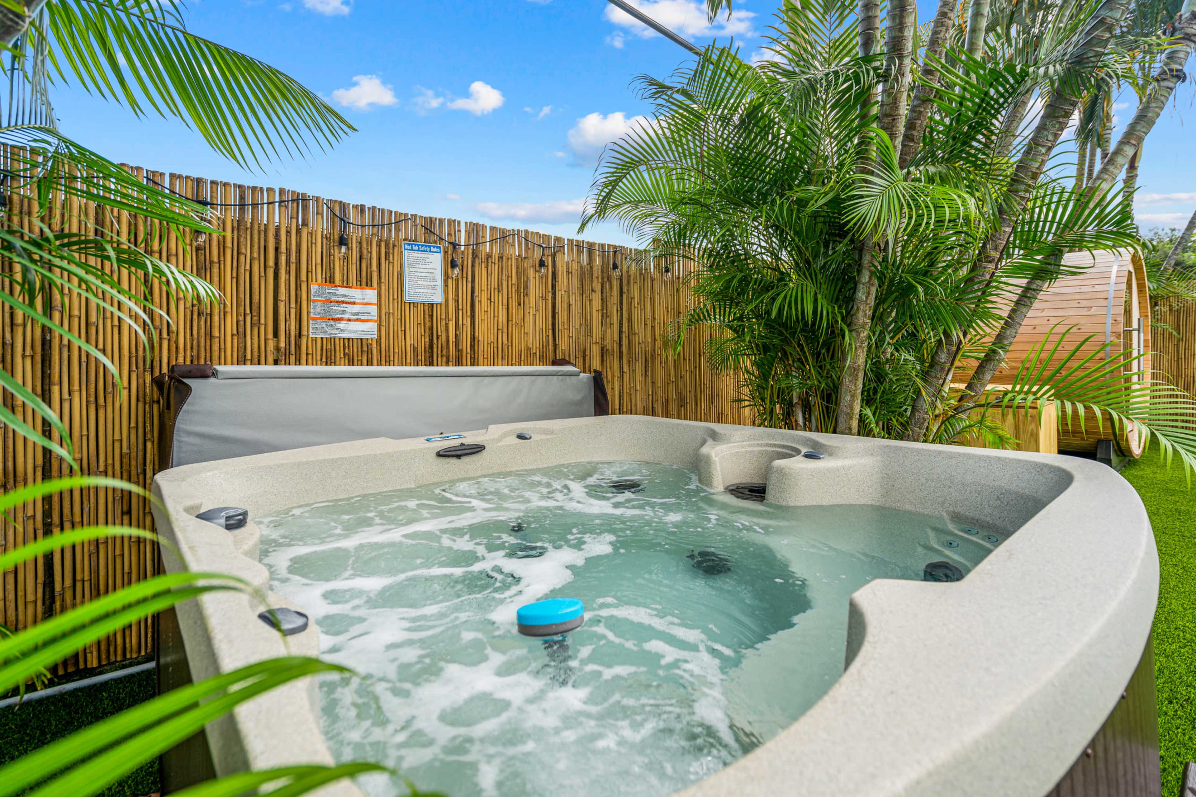 A hot tub with bubbling water is surrounded by palm plants and a bamboo fence in a backyard setting.