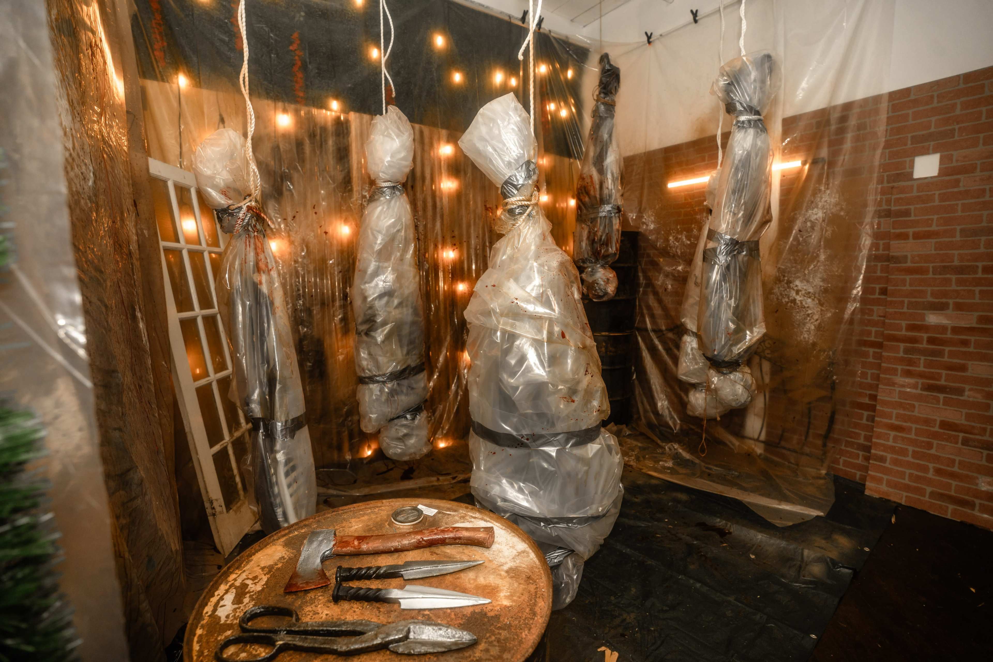 The image shows a dimly lit room with several figures wrapped in plastic and suspended from the ceiling, surrounded by tools and a table in the center.