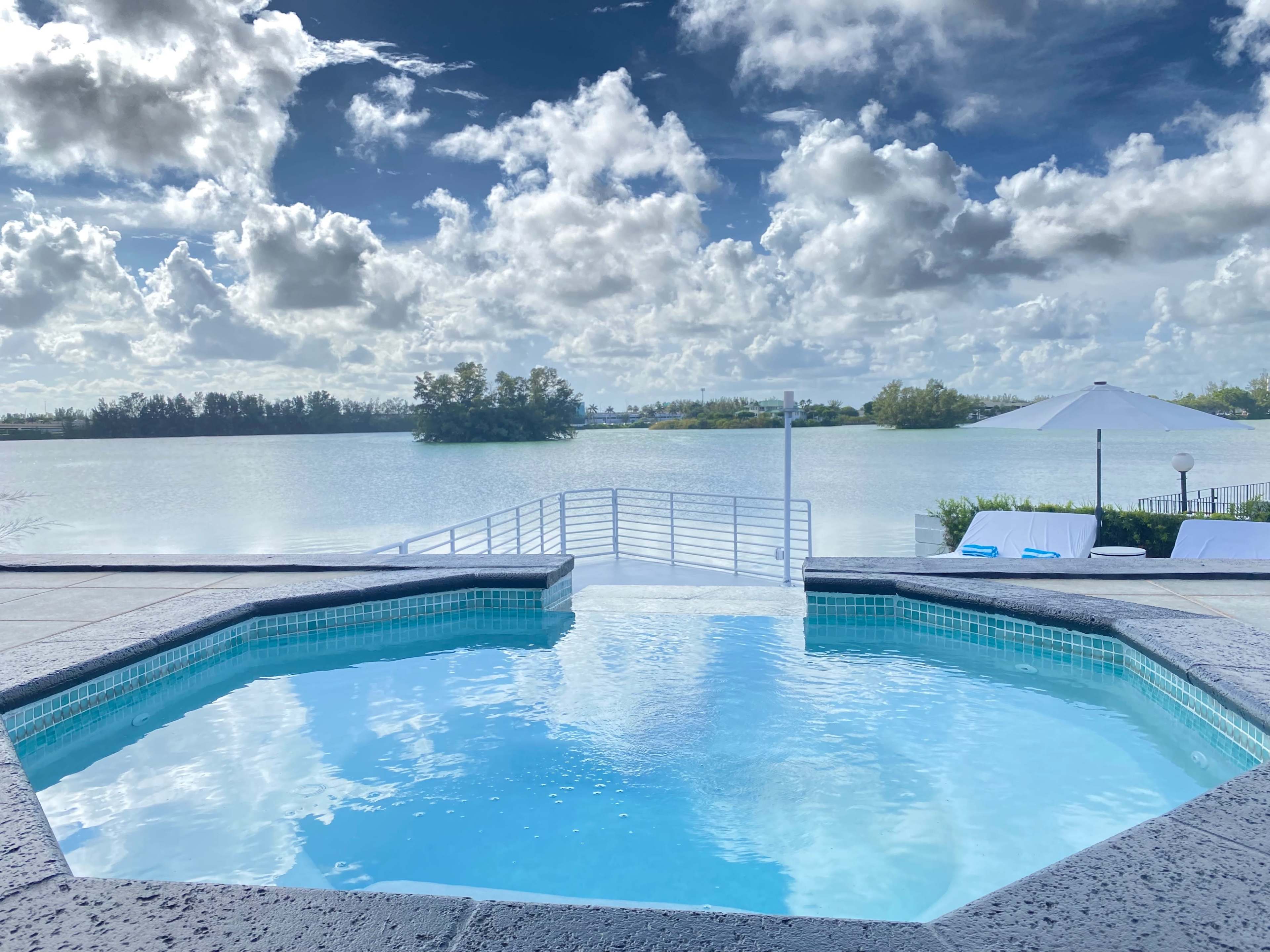 A clear swimming pool overlooks a calm body of water surrounded by lush greenery and scattered clouds in the sky.