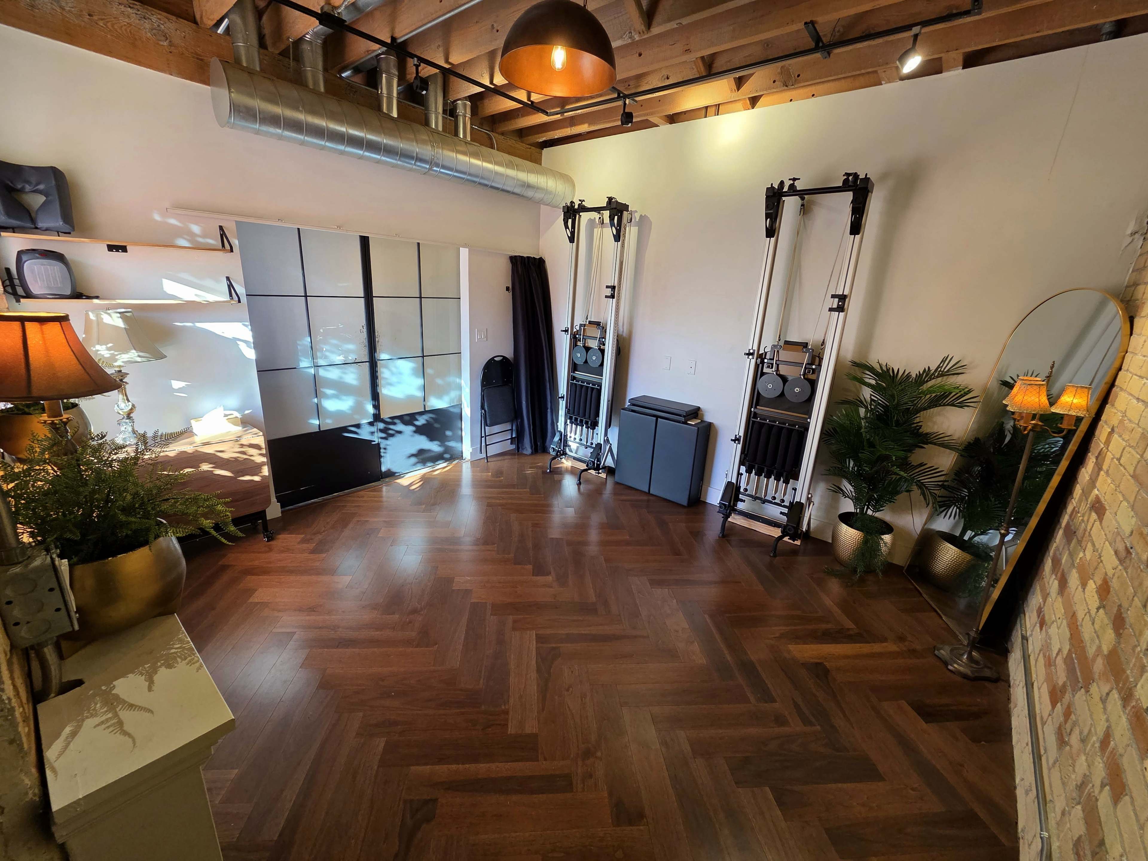 The image shows a well-lit room featuring a wooden floor, exercise equipment, a large mirror, and a mix of decorative plants and lighting elements.