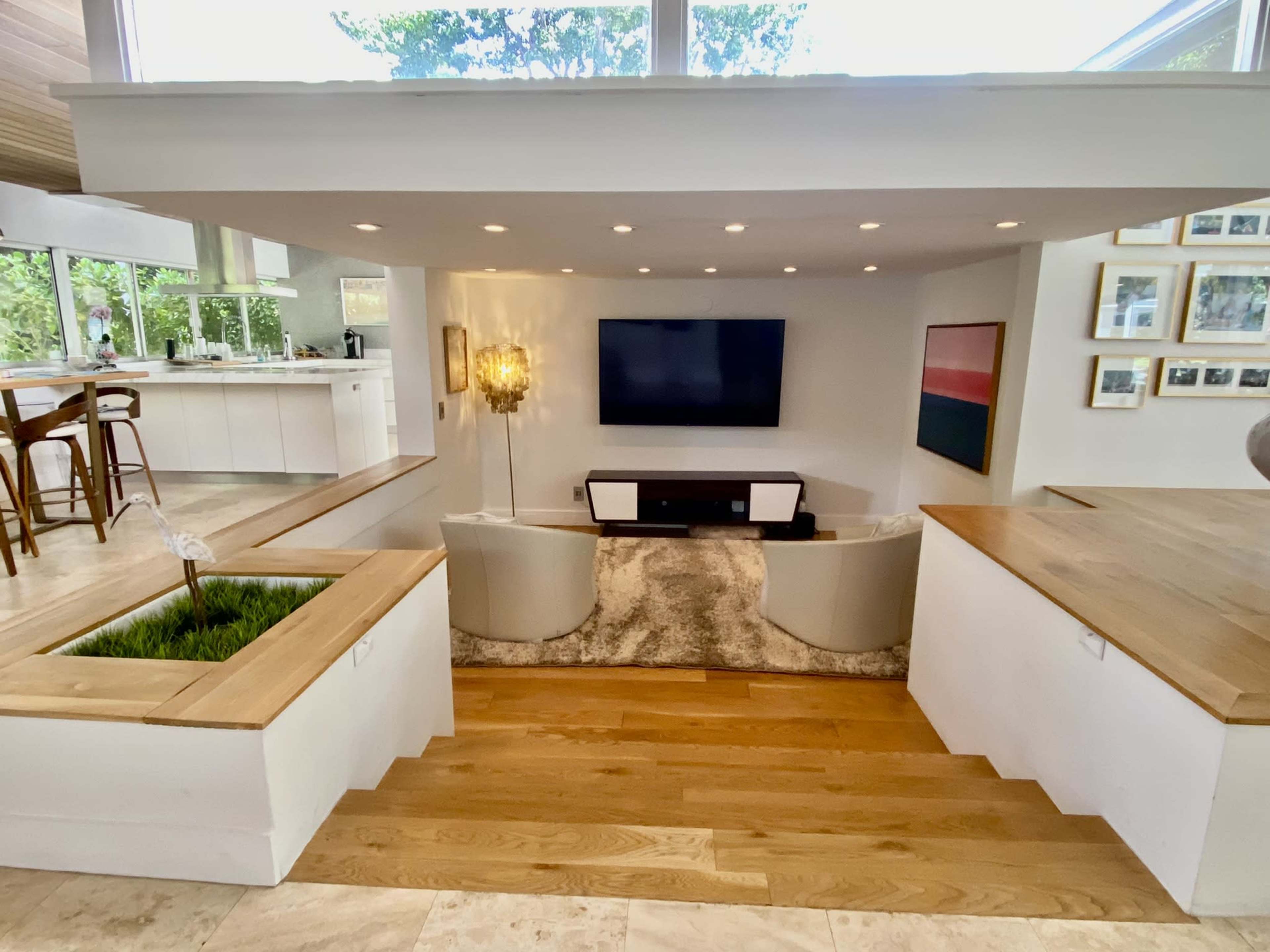 A living room features a sunken seating area with two chairs and a television on the wall, overlooking a kitchen area and illuminated by natural light from large windows.