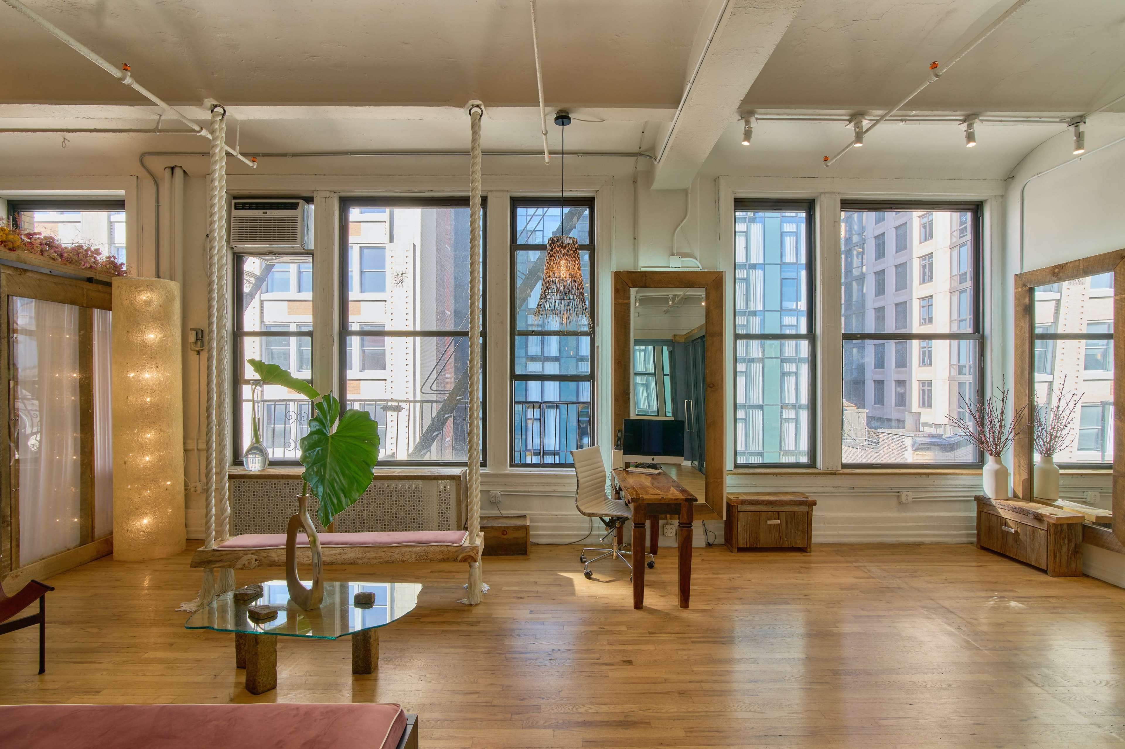 The image shows a bright room with large windows, hardwood floors, and minimal furniture, including a glass table, a wooden desk, and a seating area.