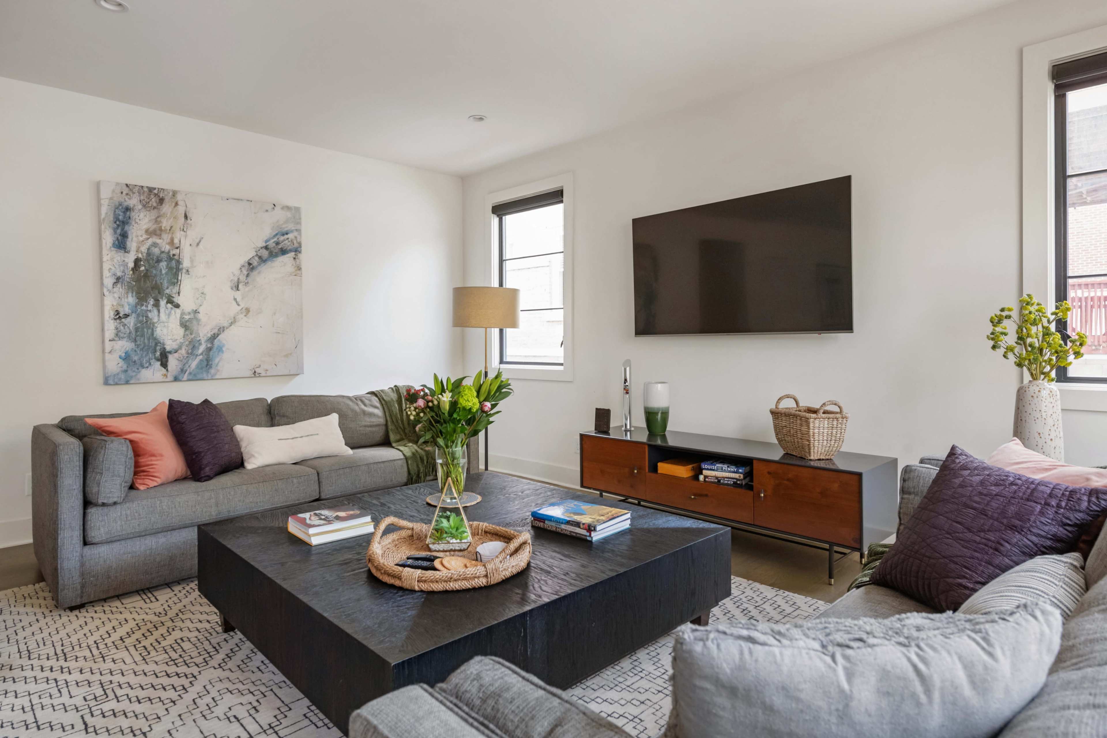 The image shows a modern living room with two gray sofas, a large black coffee table, a flat-screen television mounted on the wall, and decorative plants.