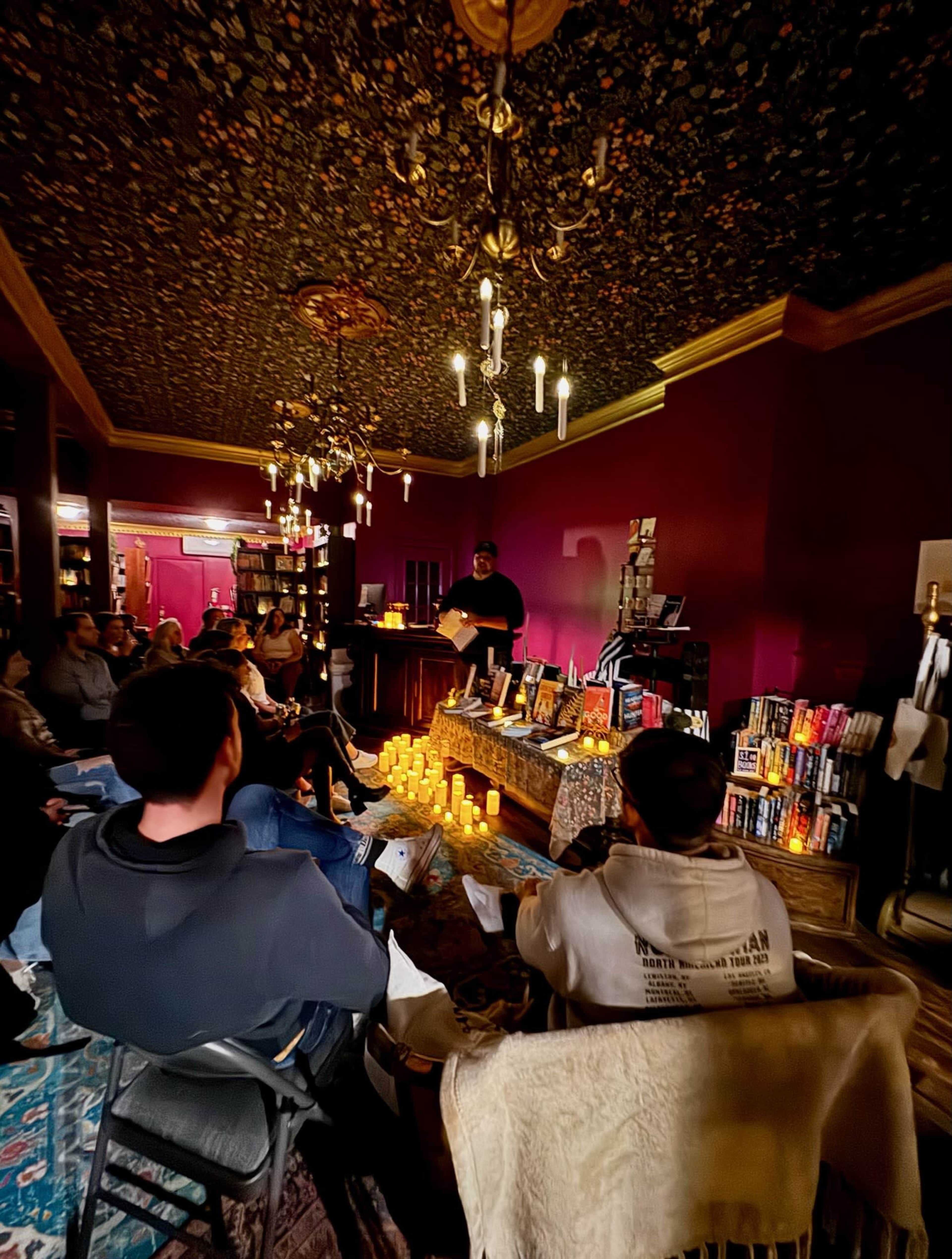 A person stands at a reading table in a candlelit room filled with shelves of books, while an audience sits in chairs arranged towards the front.