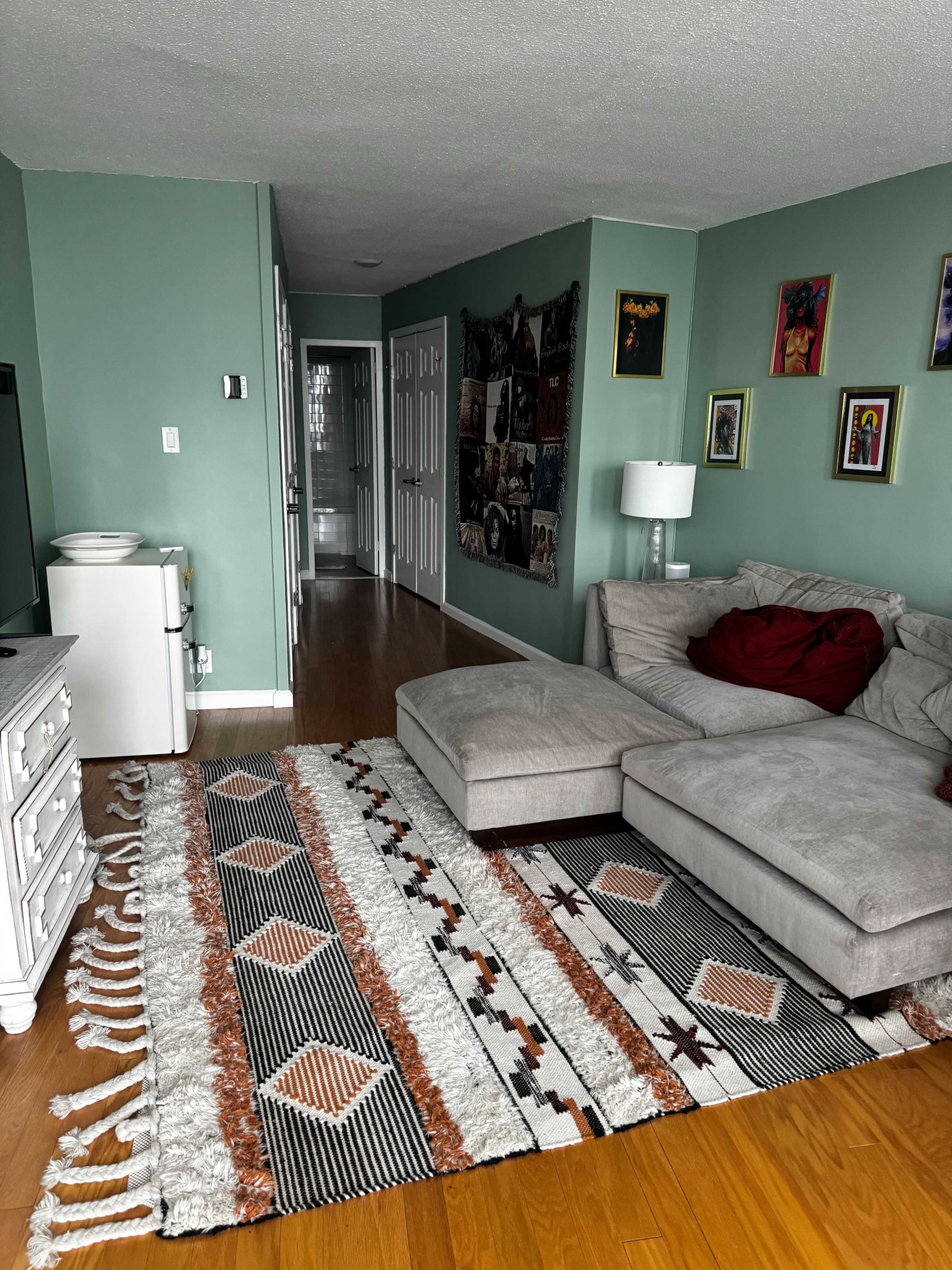 The image shows a living room with a sectional couch, a decorative area rug, a small fridge, and a wall decorated with framed pictures.