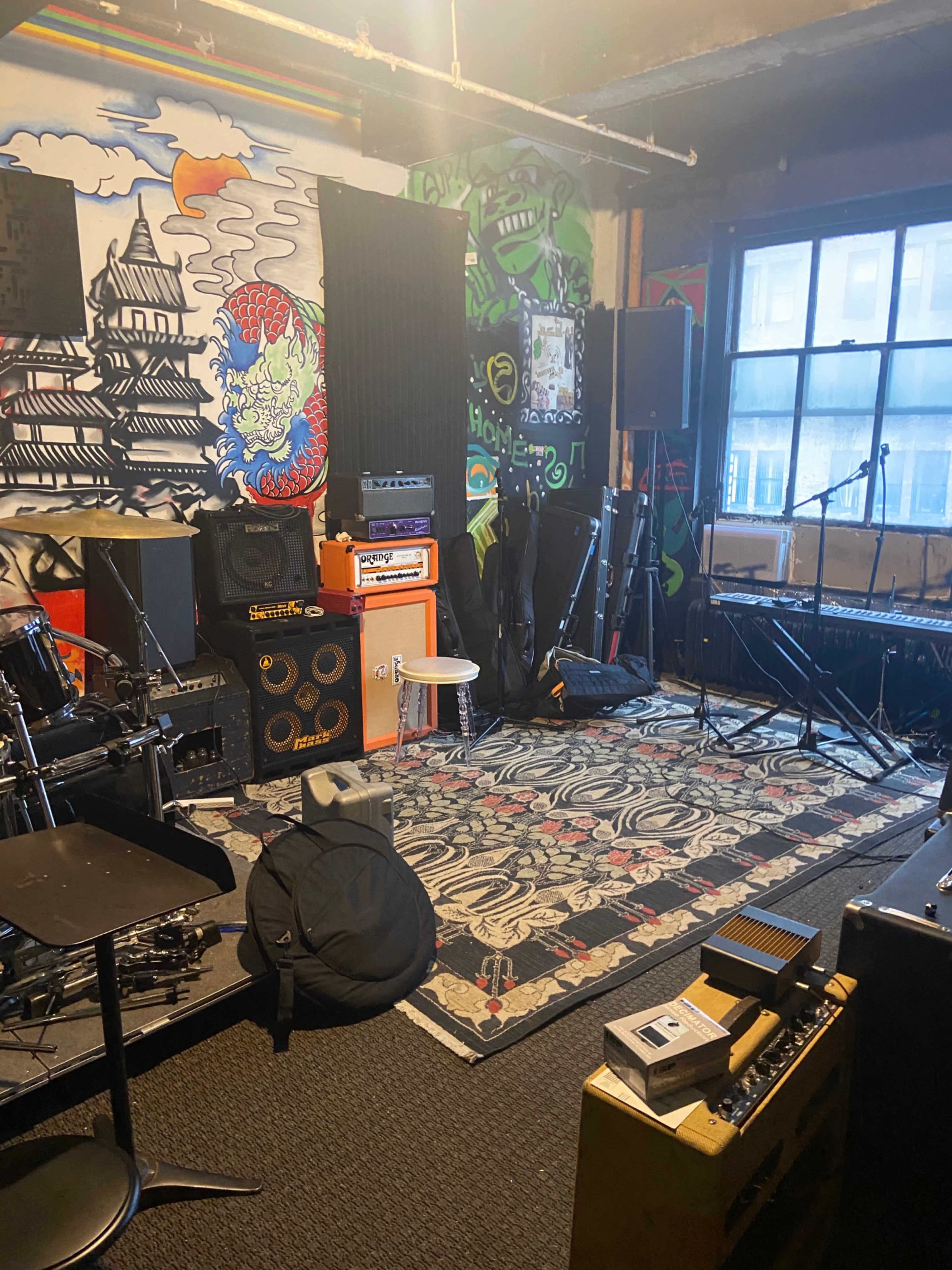 The image shows a rehearsal space decorated with a vibrant mural and equipped with various musical instruments, amplifiers, and sound equipment.