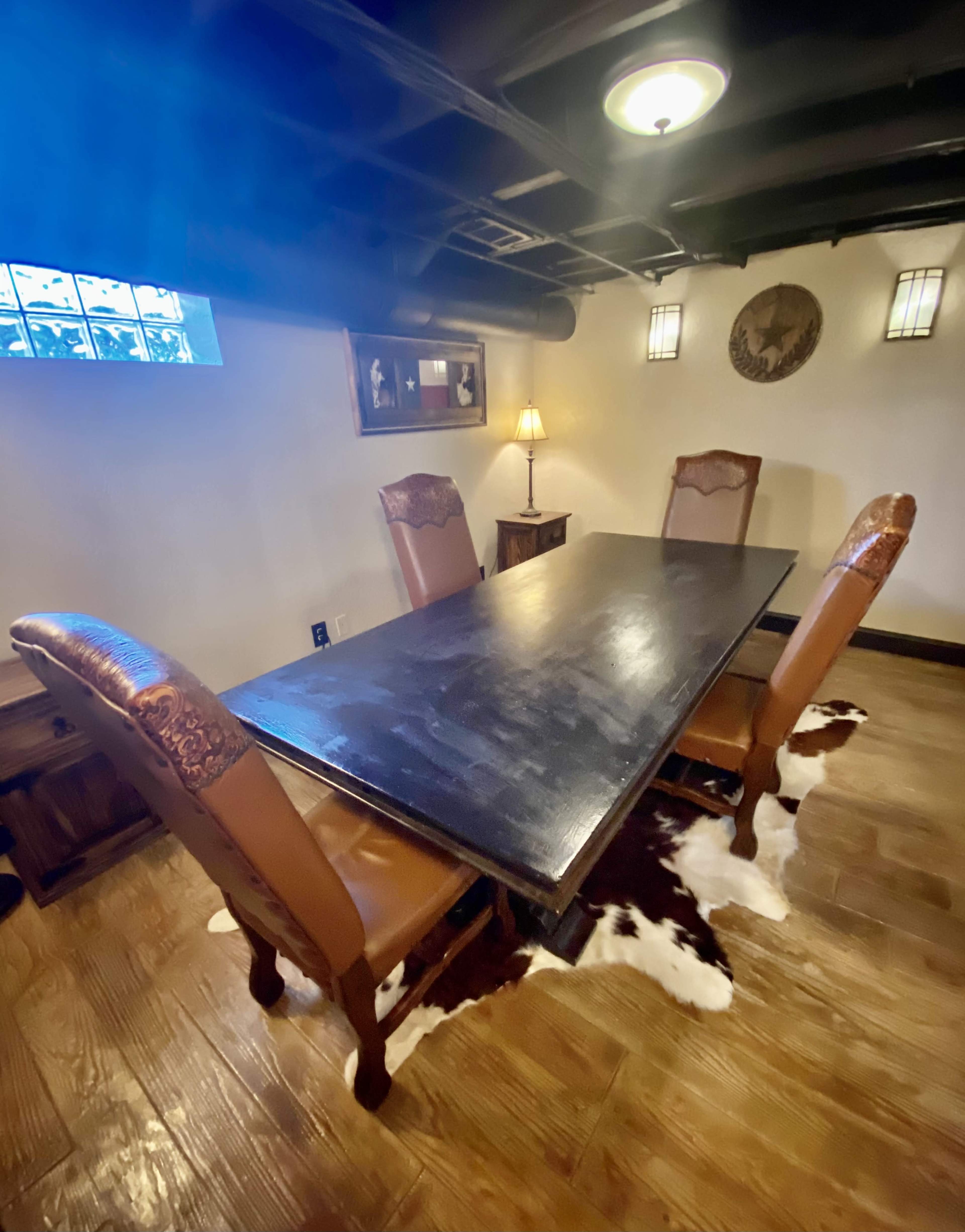 A long, dark wooden table is surrounded by three chairs on a cowhide rug in a well-lit basement room.