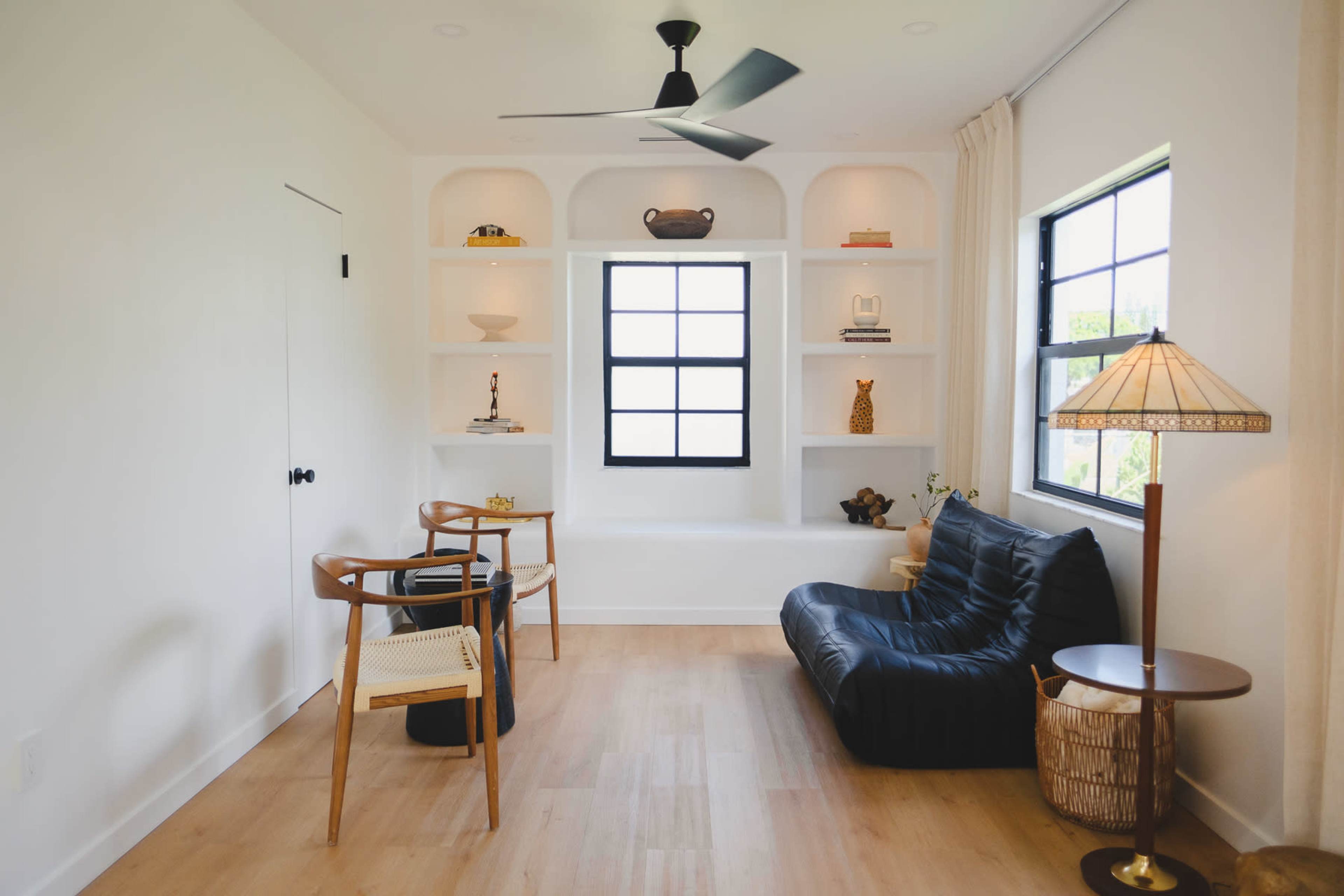 The image shows a modern room with light wood flooring, a black leather chair, a small round table, and built-in shelves displaying various decorative items.