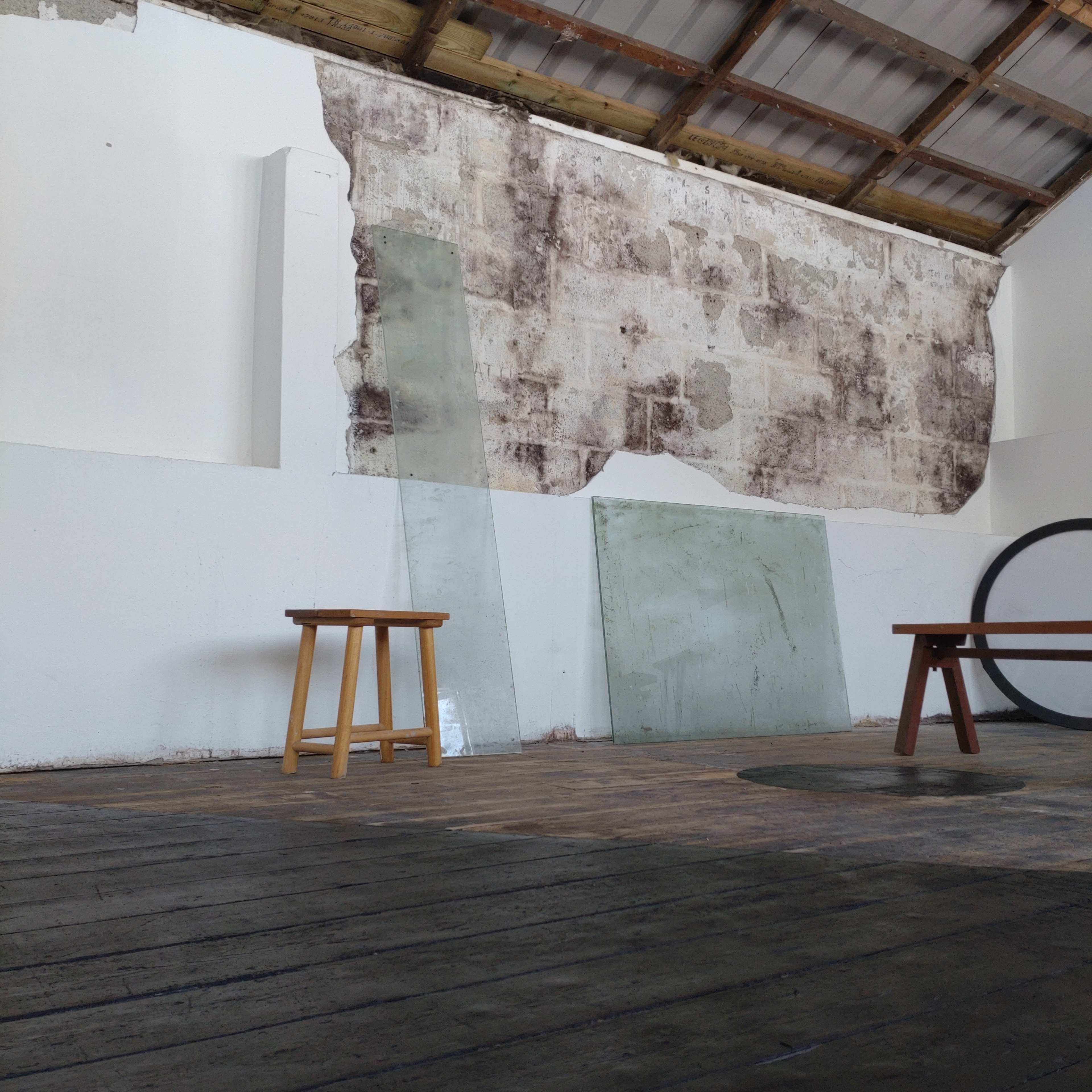 A wooden stool in the foreground, with two large glass panels leaning against a textured, partially exposed wall and a circular object on the floor.