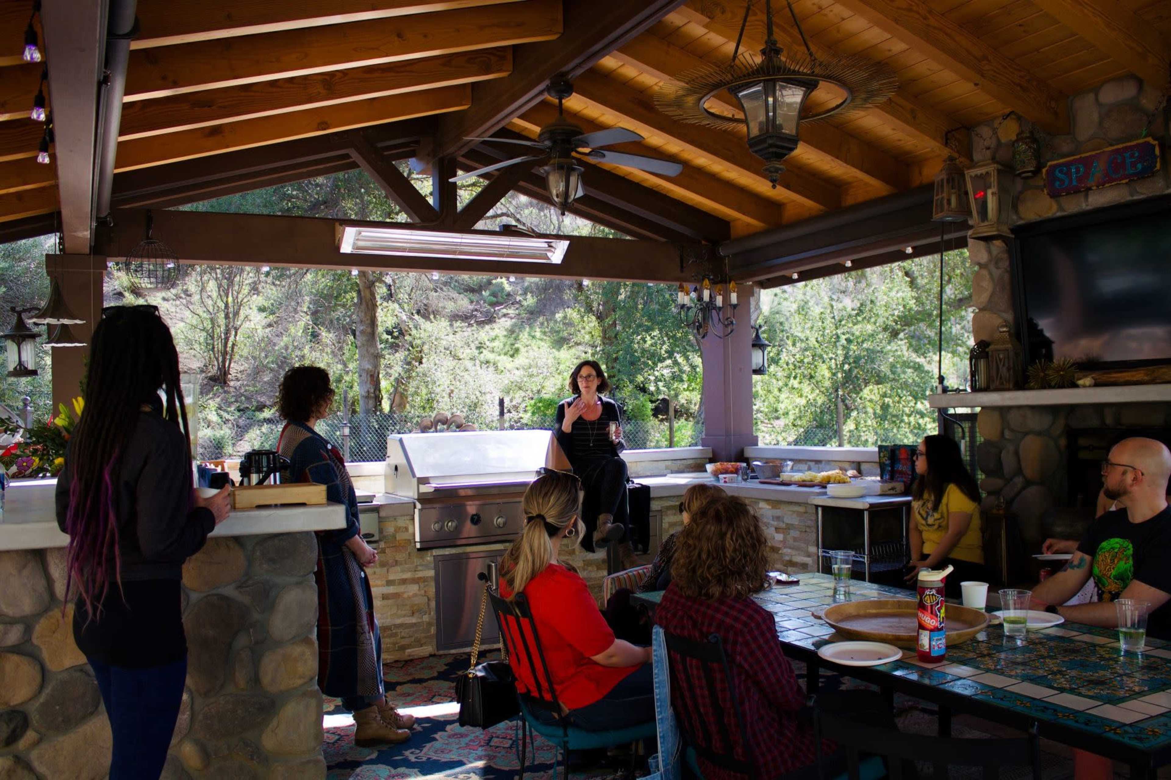 A group of people is gathered in an outdoor kitchen area, with one person speaking as others listen attentively.