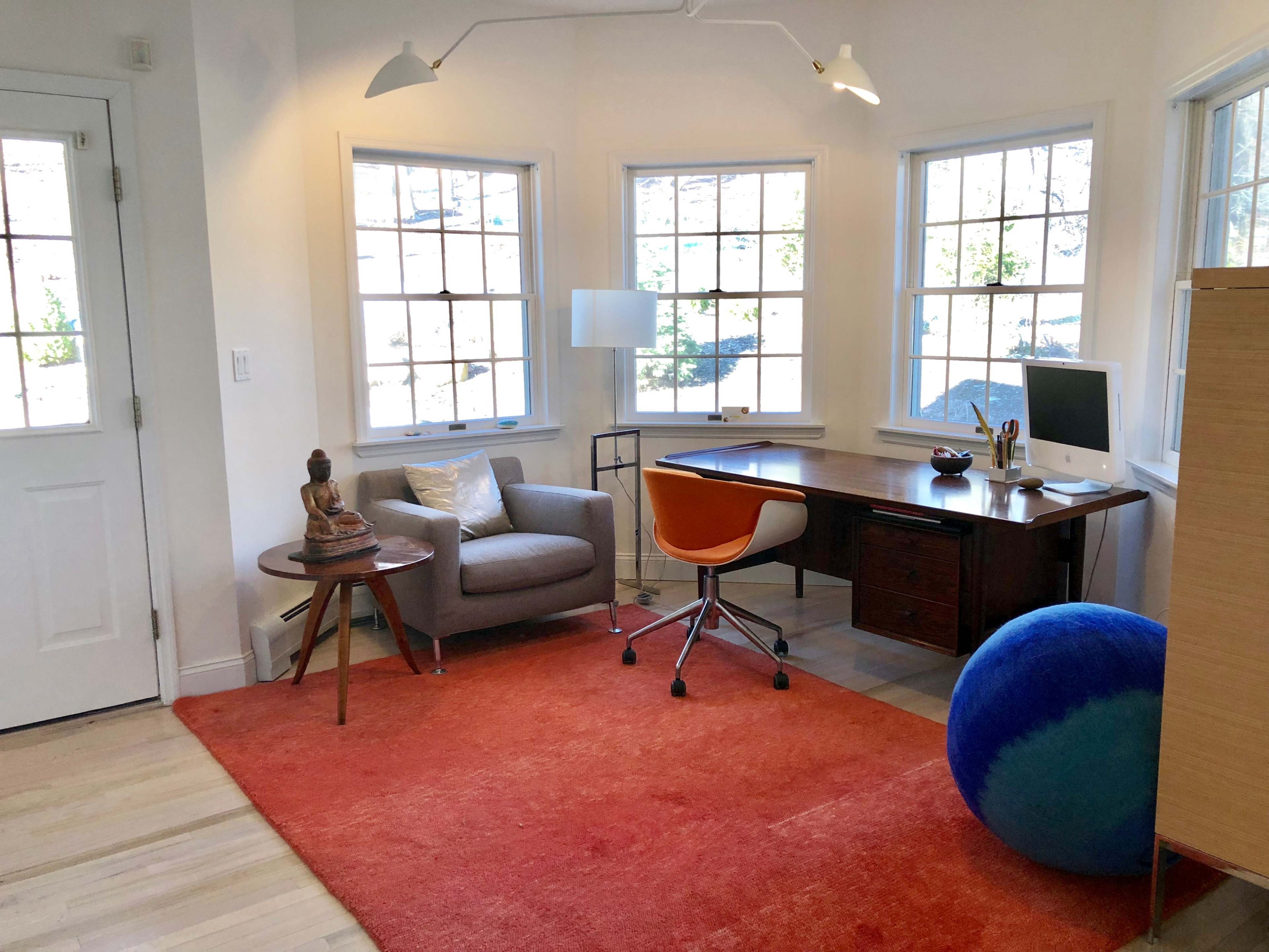 A bright home office features a wooden desk with an iMac, an orange chair, a gray armchair, a blue exercise ball, and large windows letting in natural light.