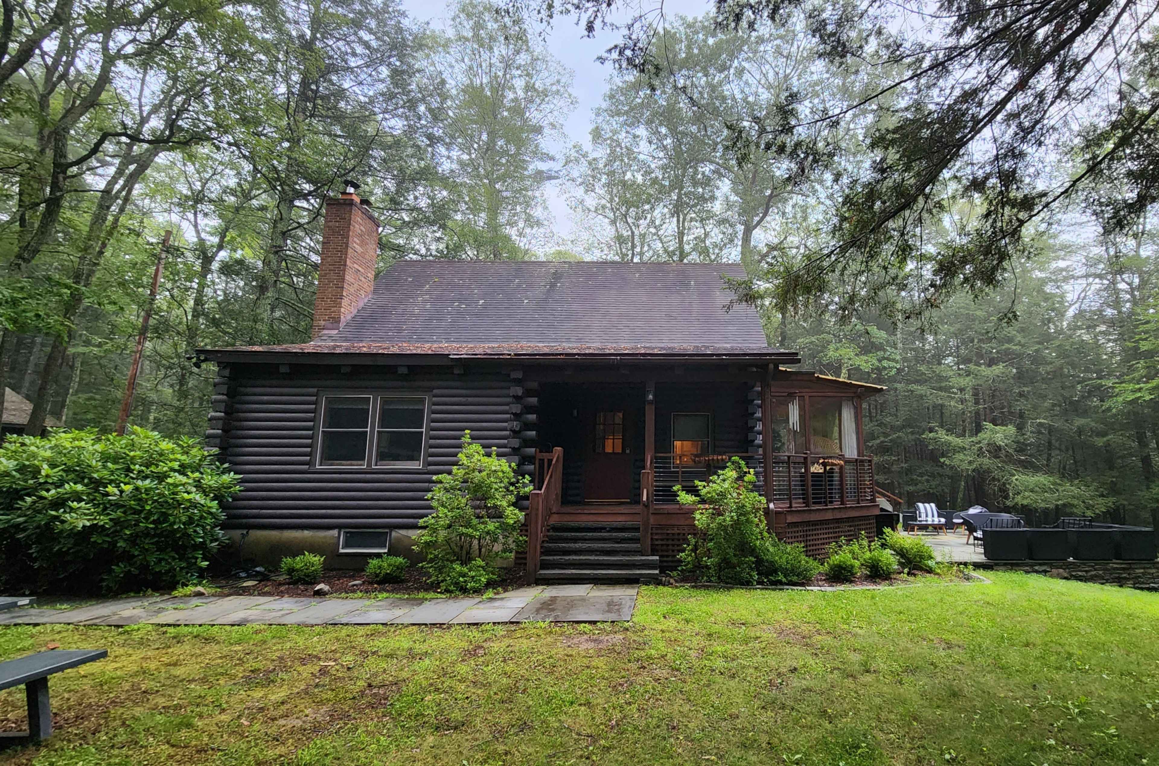 A log cabin is situated in a wooded area with green grass and stone walkways leading to its entrance.