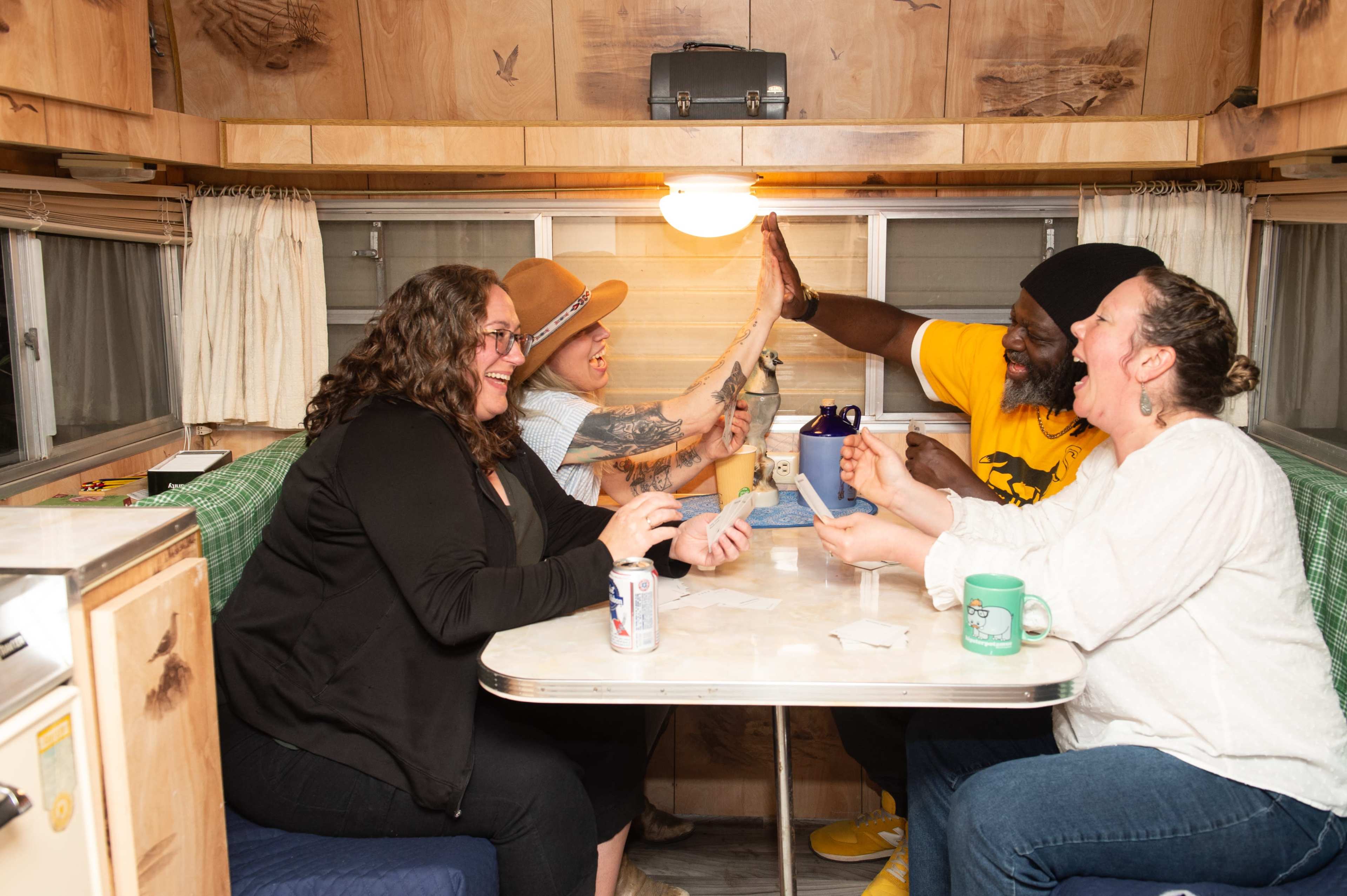 Four people are seated around a table in a retro camper, engaging in a lively game or activity while enjoying drinks.