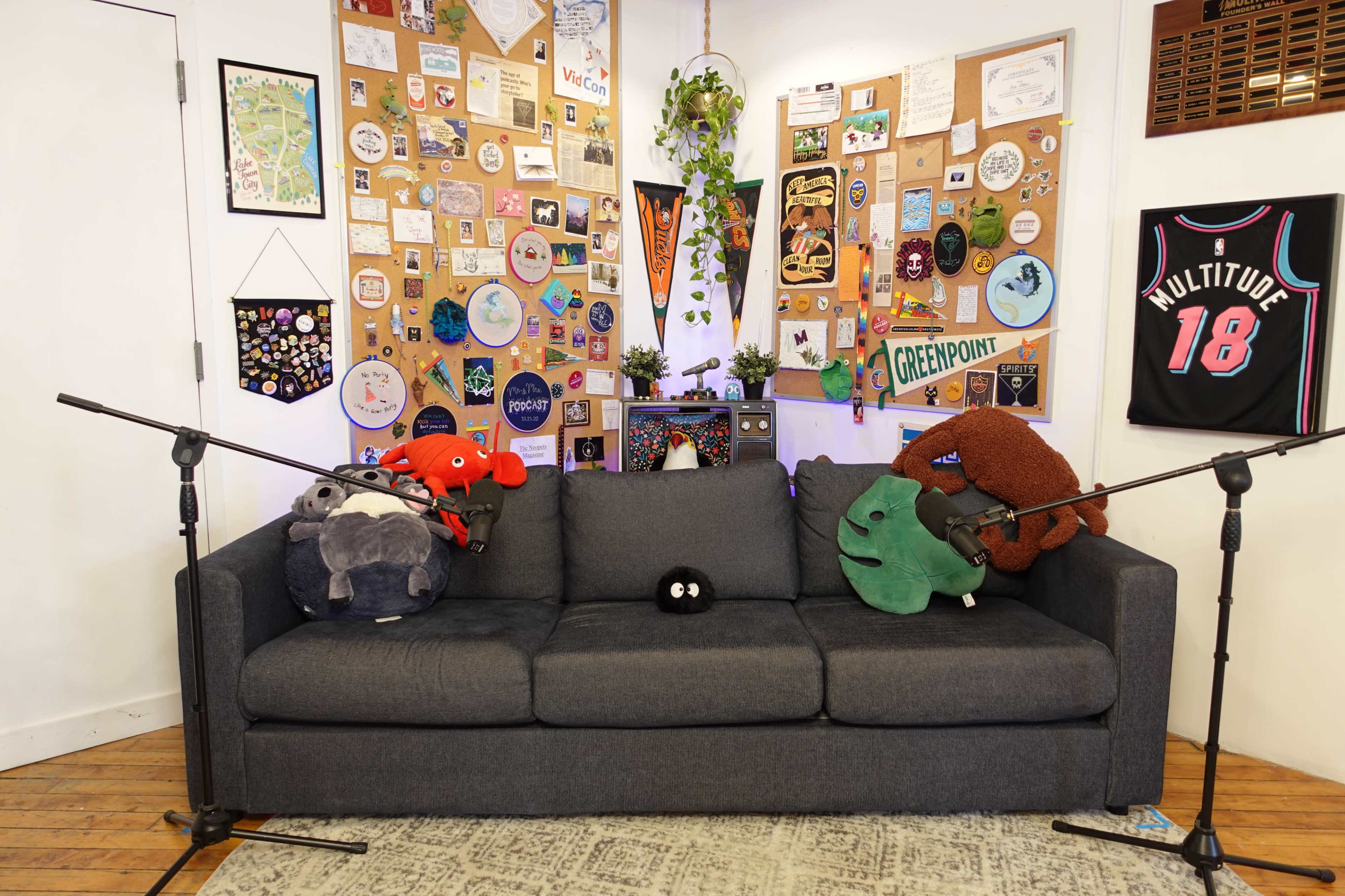 A gray sofa adorned with various plush toys, positioned in front of a wall covered with stickers and memorabilia, and flanked by two microphone stands.