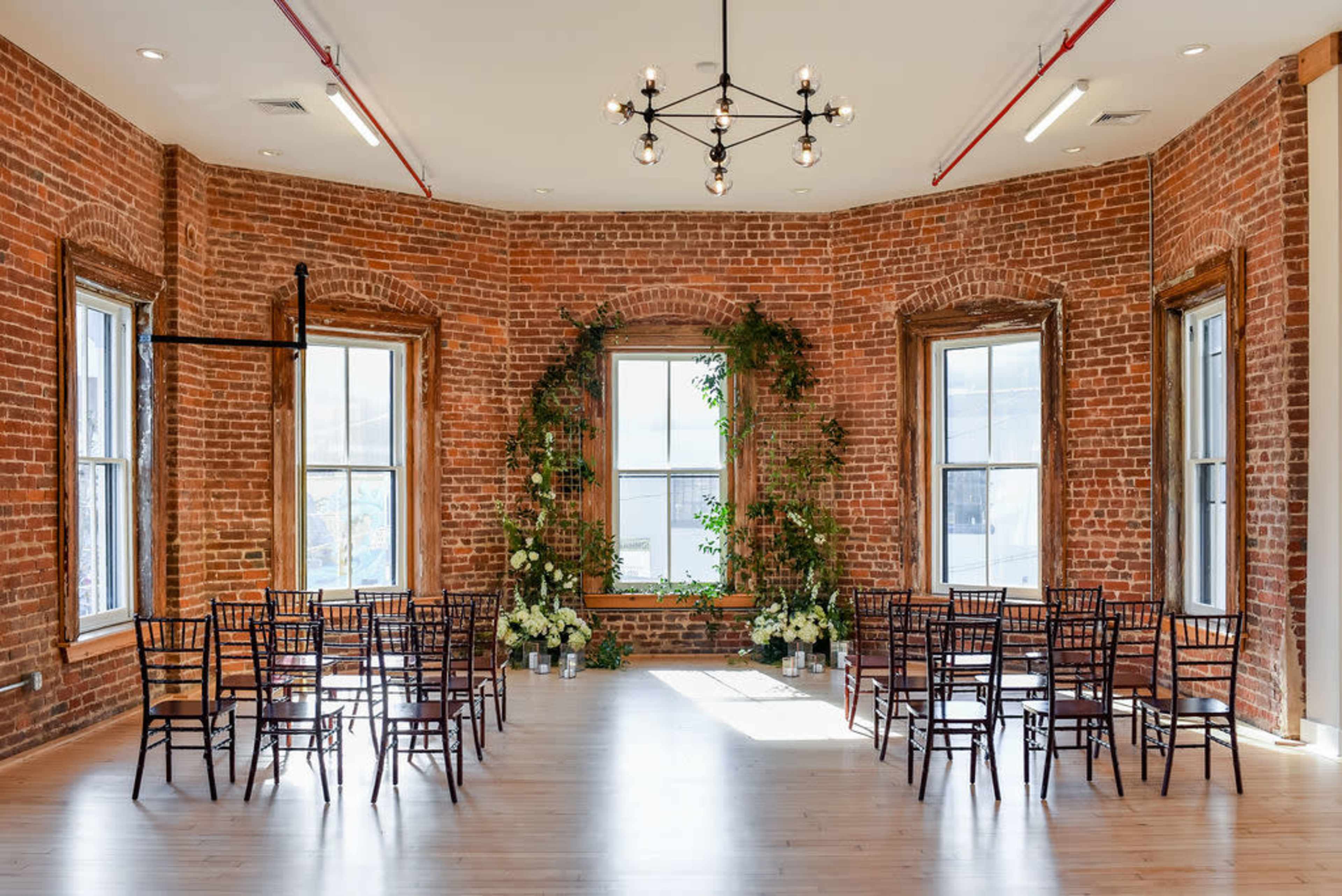 A room with exposed brick walls, adorned with plants and floral arrangements, featuring a circular seating arrangement of dark wooden chairs.