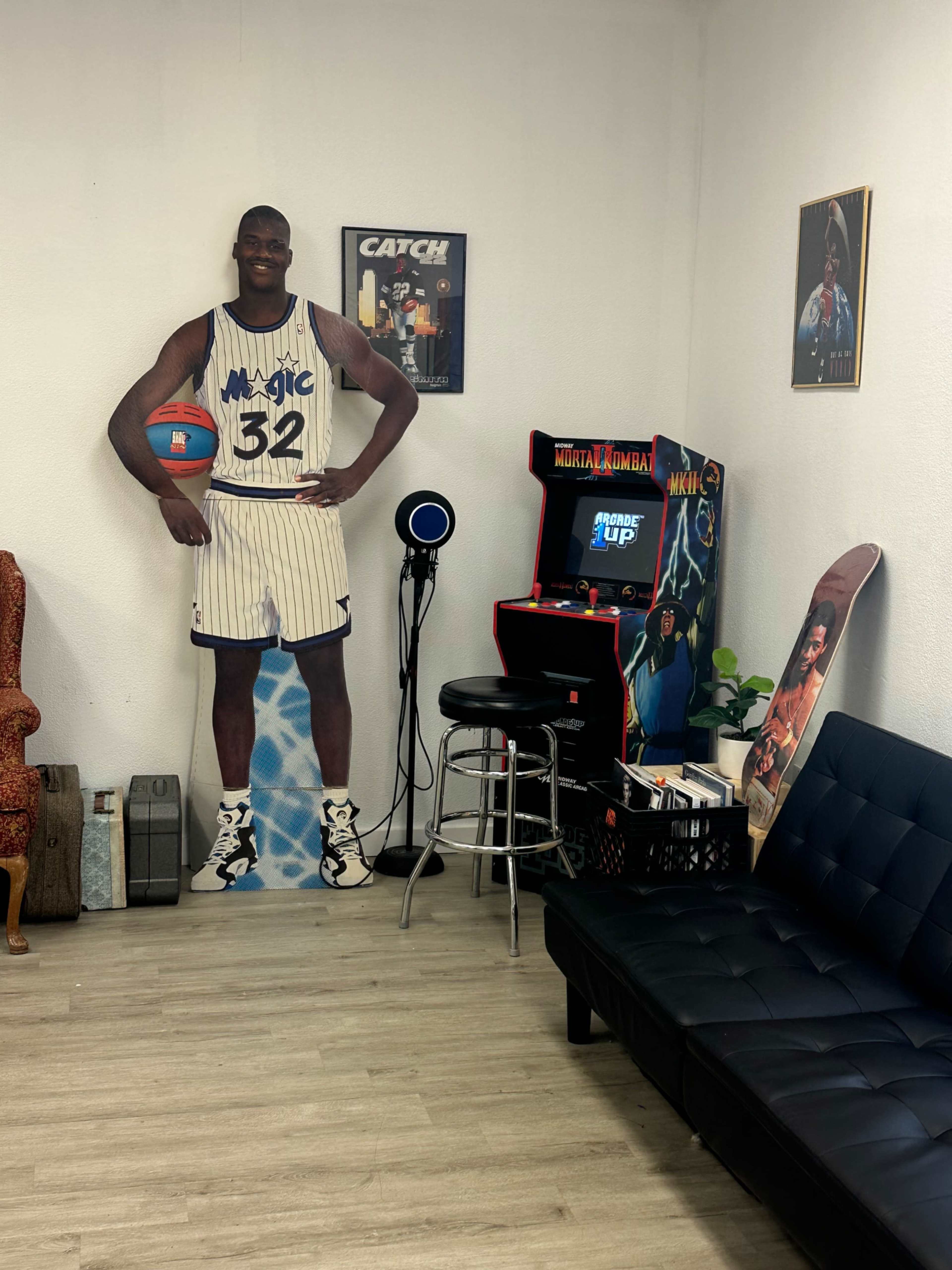 The scene features a life-sized cardboard cutout of a basketball player next to a vintage arcade machine in a recreational room with seating.