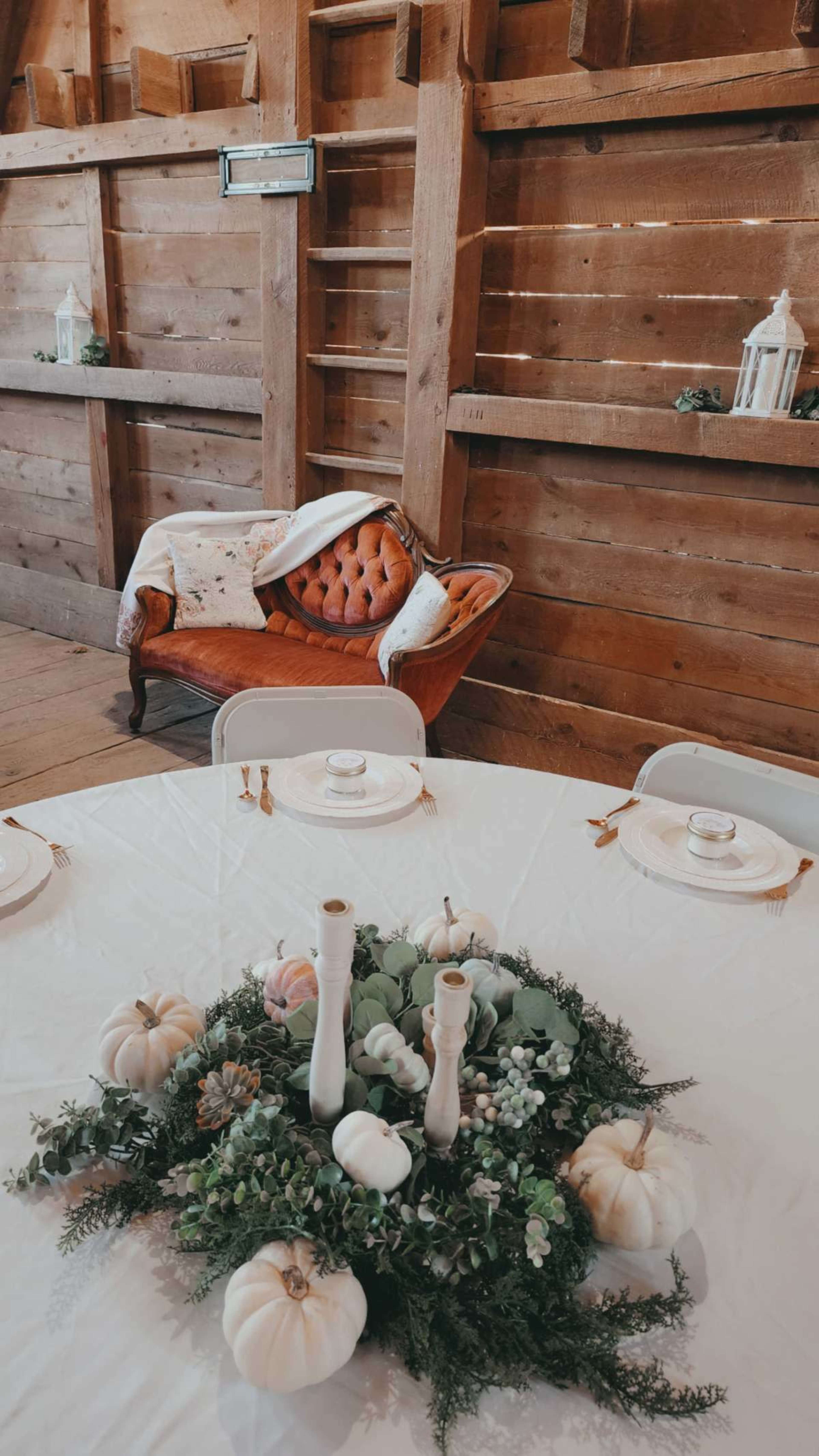 A round table with a centerpiece of greenery and small white pumpkins is set in a rustic barn, featuring a vintage orange couch in the background.
