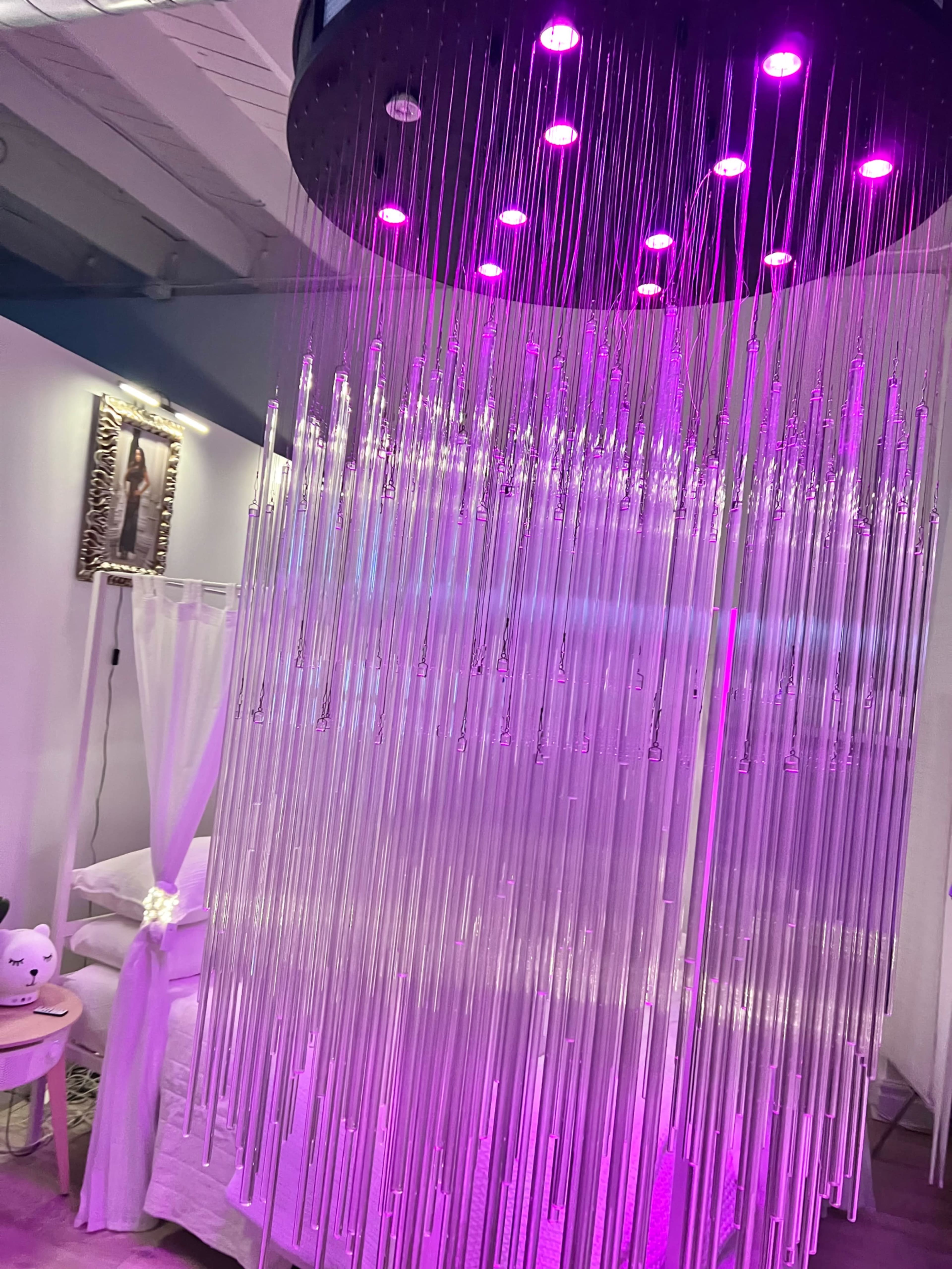The image shows a decorative installation featuring hanging clear tubes illuminated by purple lights, set in a room with white furnishings and a canopy bed.