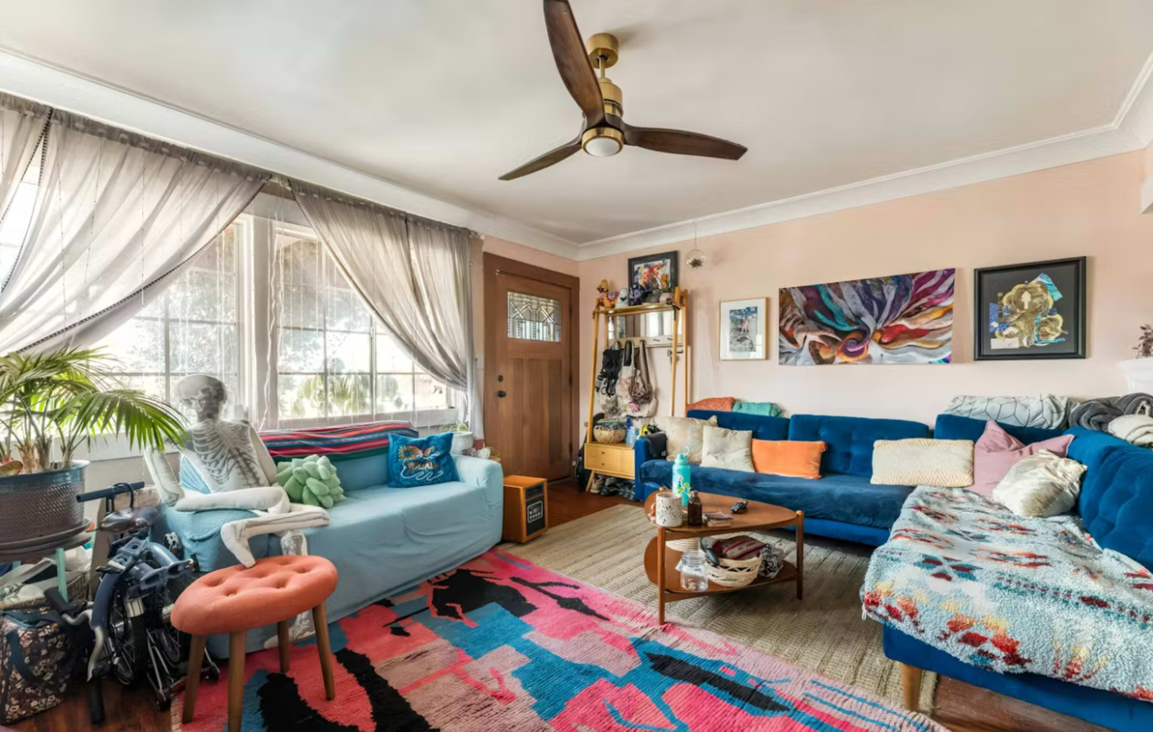 The image shows a cozy living room featuring a blue sectional sofa, a decorative rug, a potted plant, and a skeleton decoration, with large windows allowing natural light to fill the space.