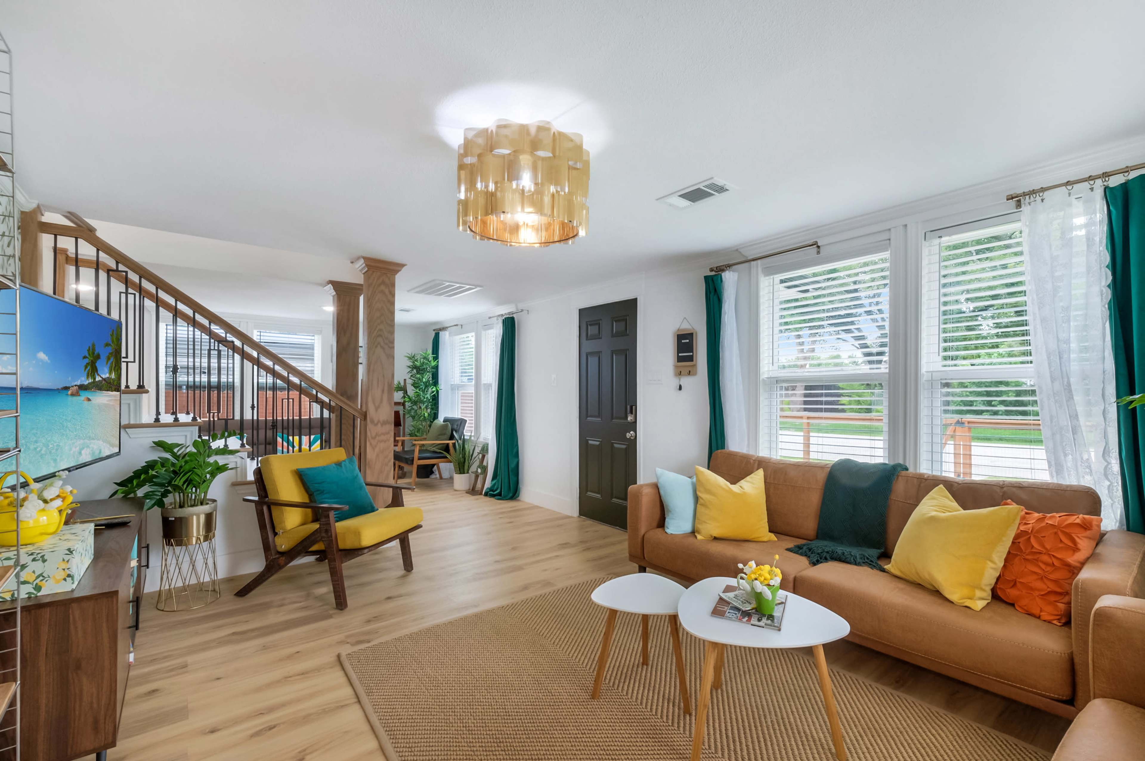 The living room features a brown sofa with colorful cushions, two small tables, and large windows with green drapes, adjacent to a staircase and a decorative TV stand.
