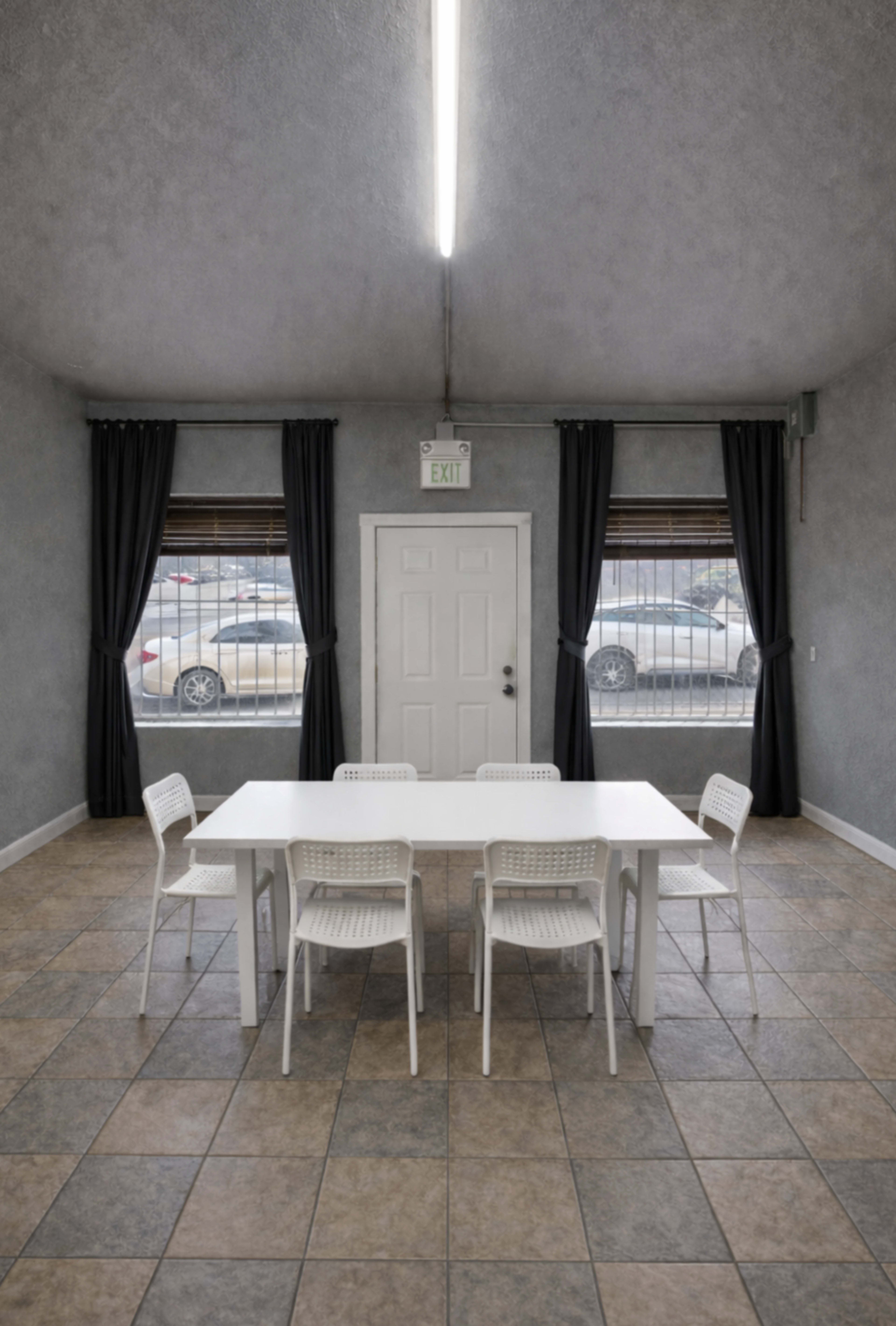 A simple, well-lit room features a white table surrounded by six chairs, with large windows dressed in dark curtains.