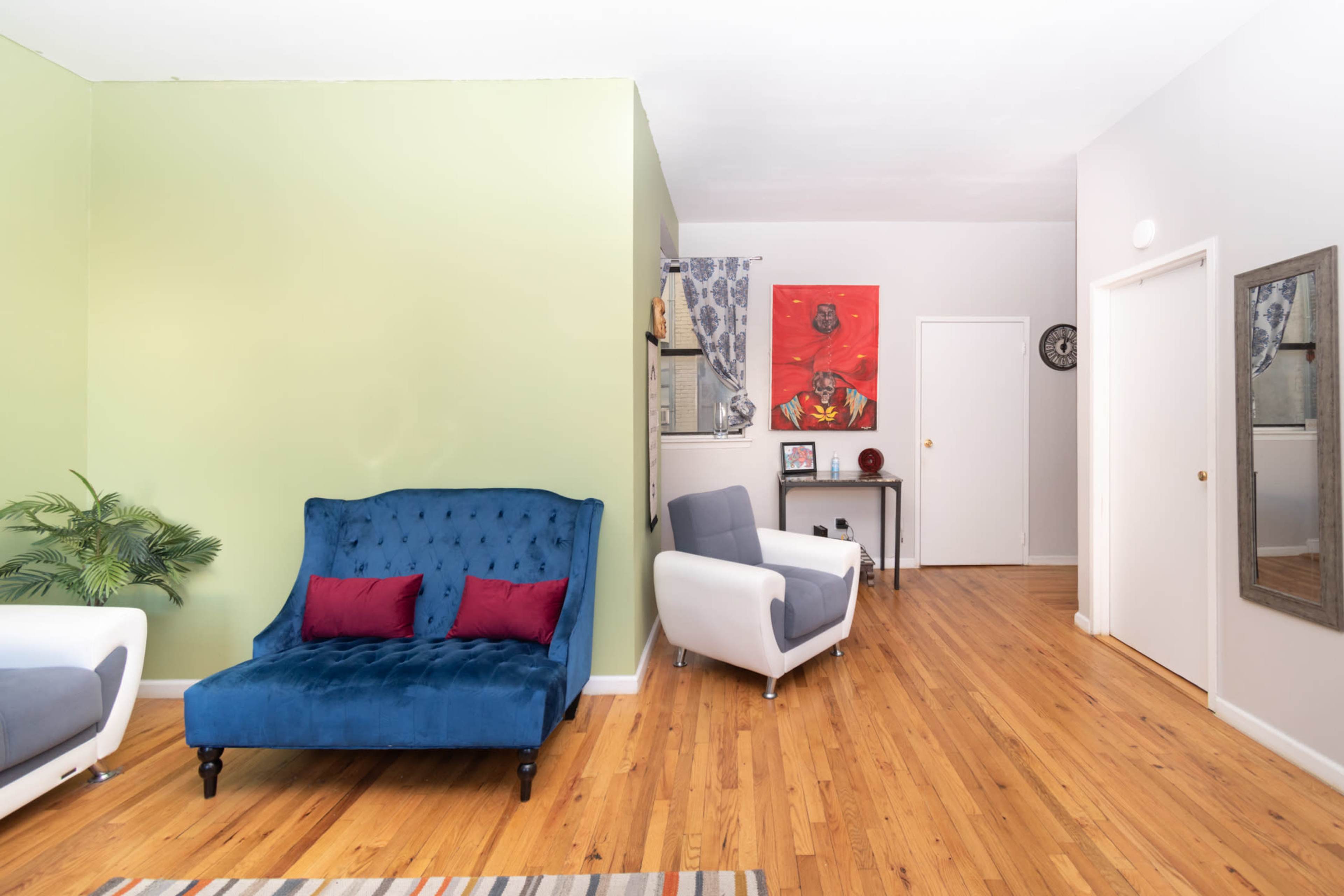 A living room featuring a blue upholstered sofa with red cushions, a small table, and light hardwood flooring, alongside a green accent wall and a doorway leading to another room.