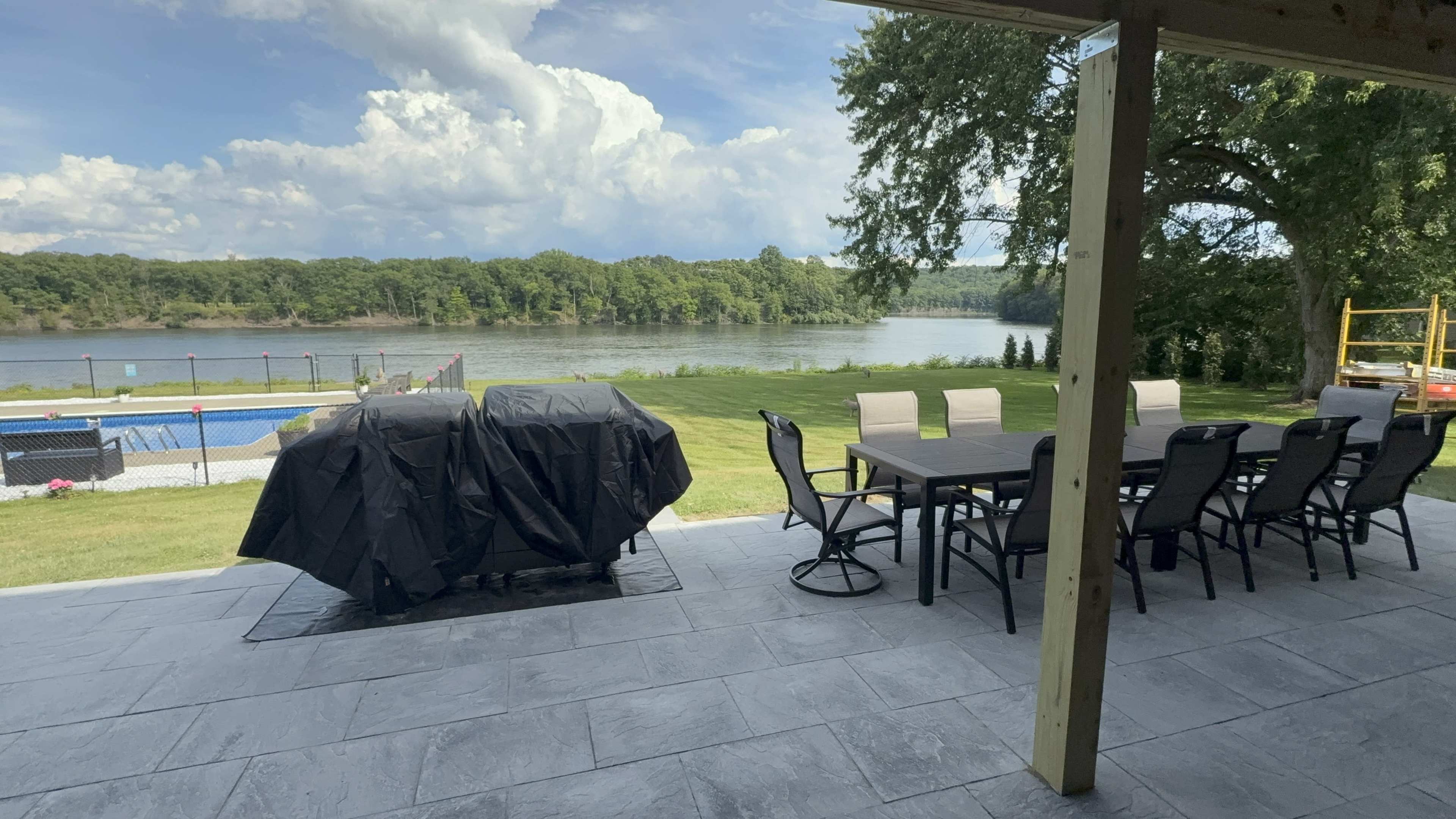 The image shows a patio area with a covered grill, a dining table with chairs, and a view of a riverbank beyond a lawn.