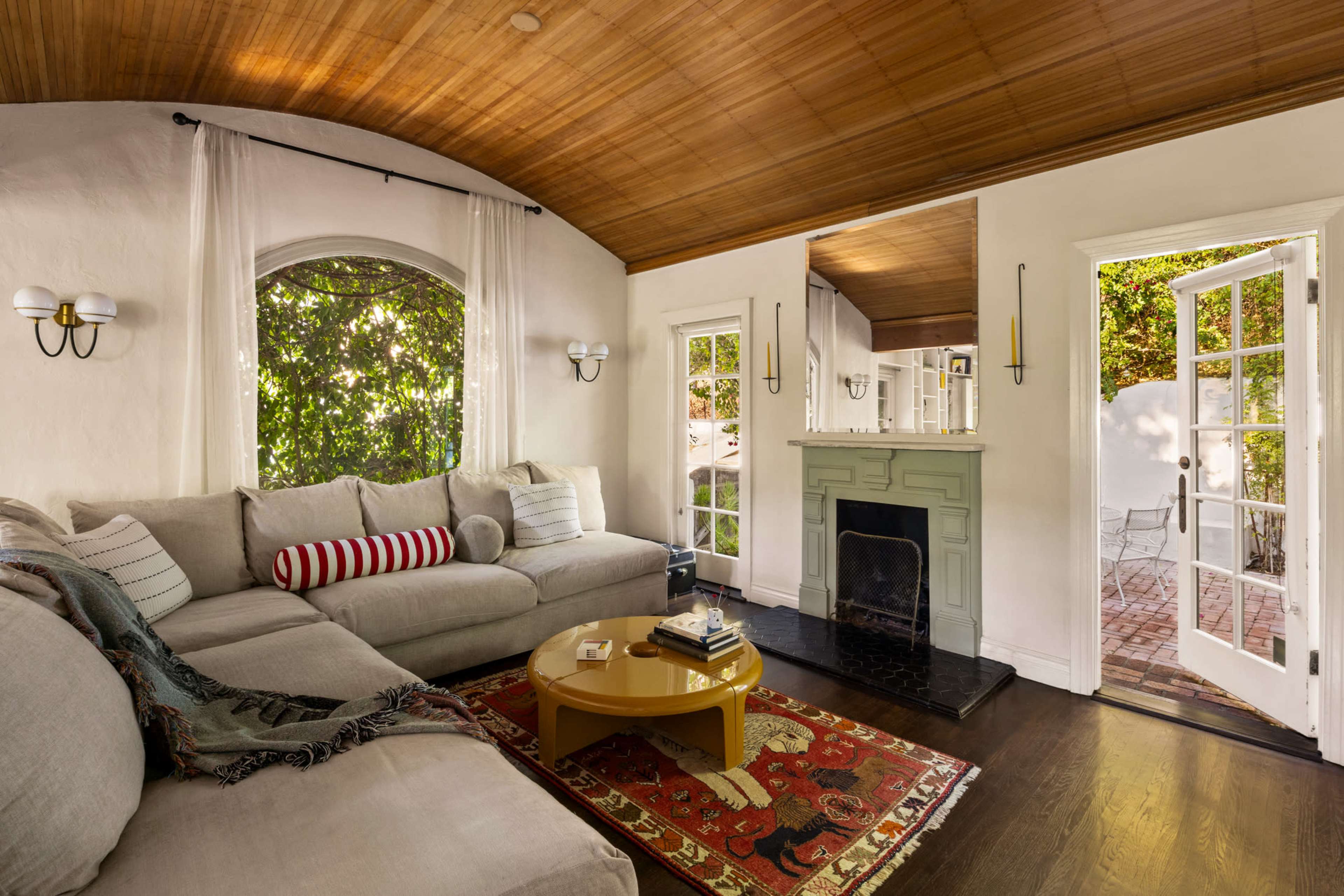 The living room features a curved wooden ceiling, large arched windows, a light-colored sectional sofa, and a fireplace, with access to an outdoor patio through French doors.