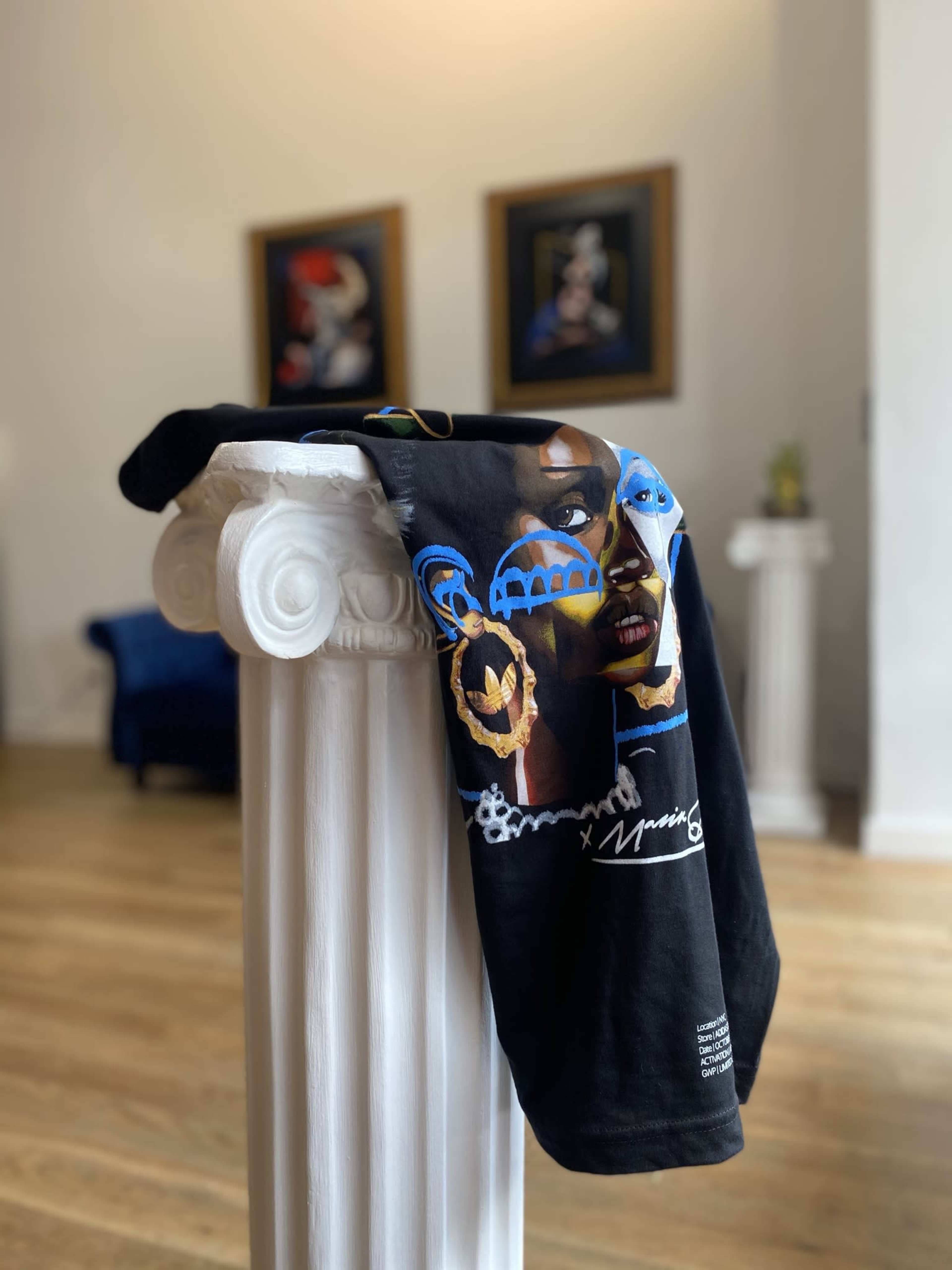 A black t-shirt with a colorful graphic design hangs over a classical white column in a well-lit room featuring artwork on the walls.