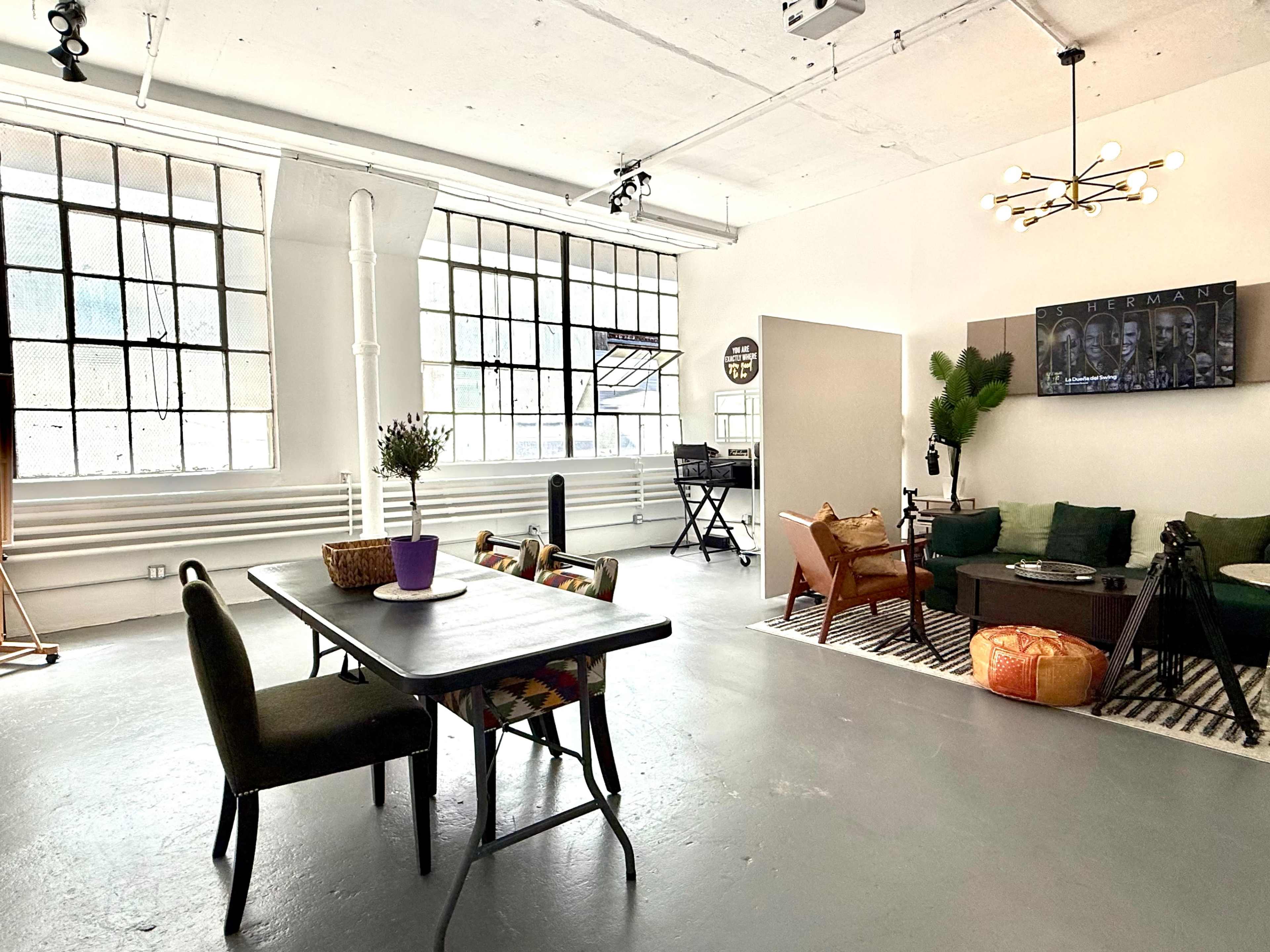 The image shows a spacious interior featuring a dining table, chairs, a living area with a sofa, and large windows allowing natural light.