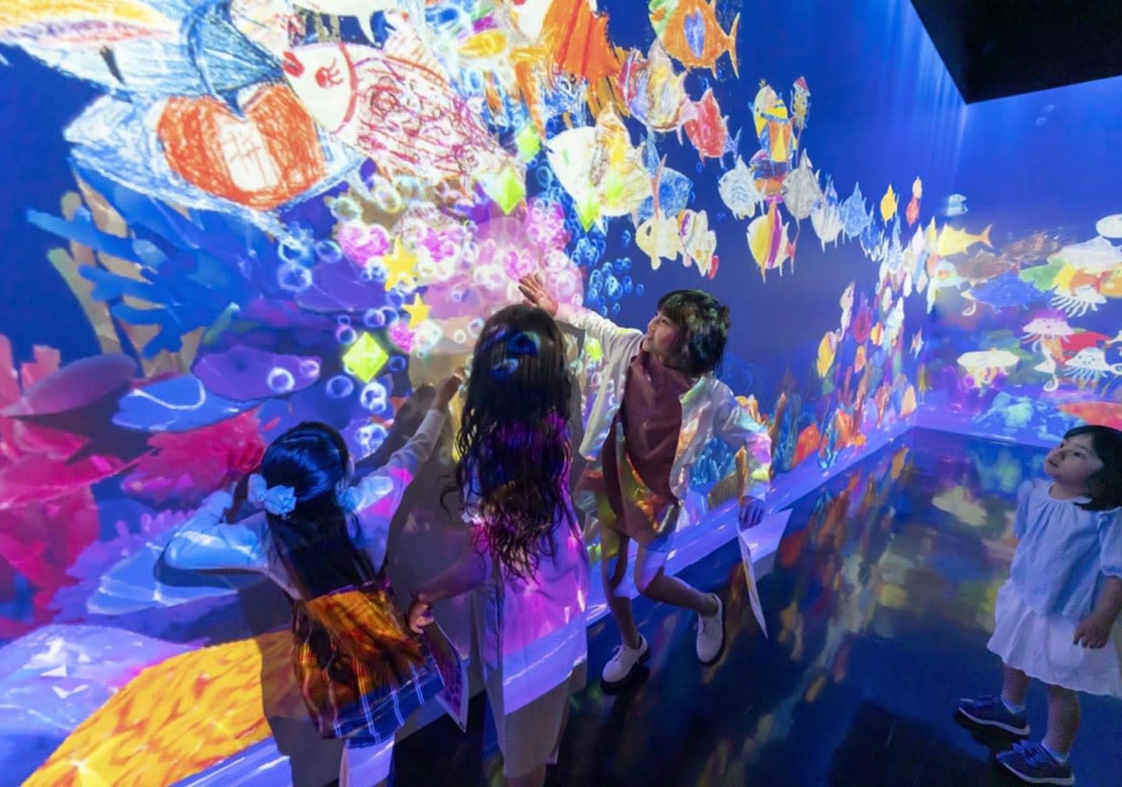 A group of children and an adult interact with a vibrant, projected underwater scene covered in colorful fish and bubbles.