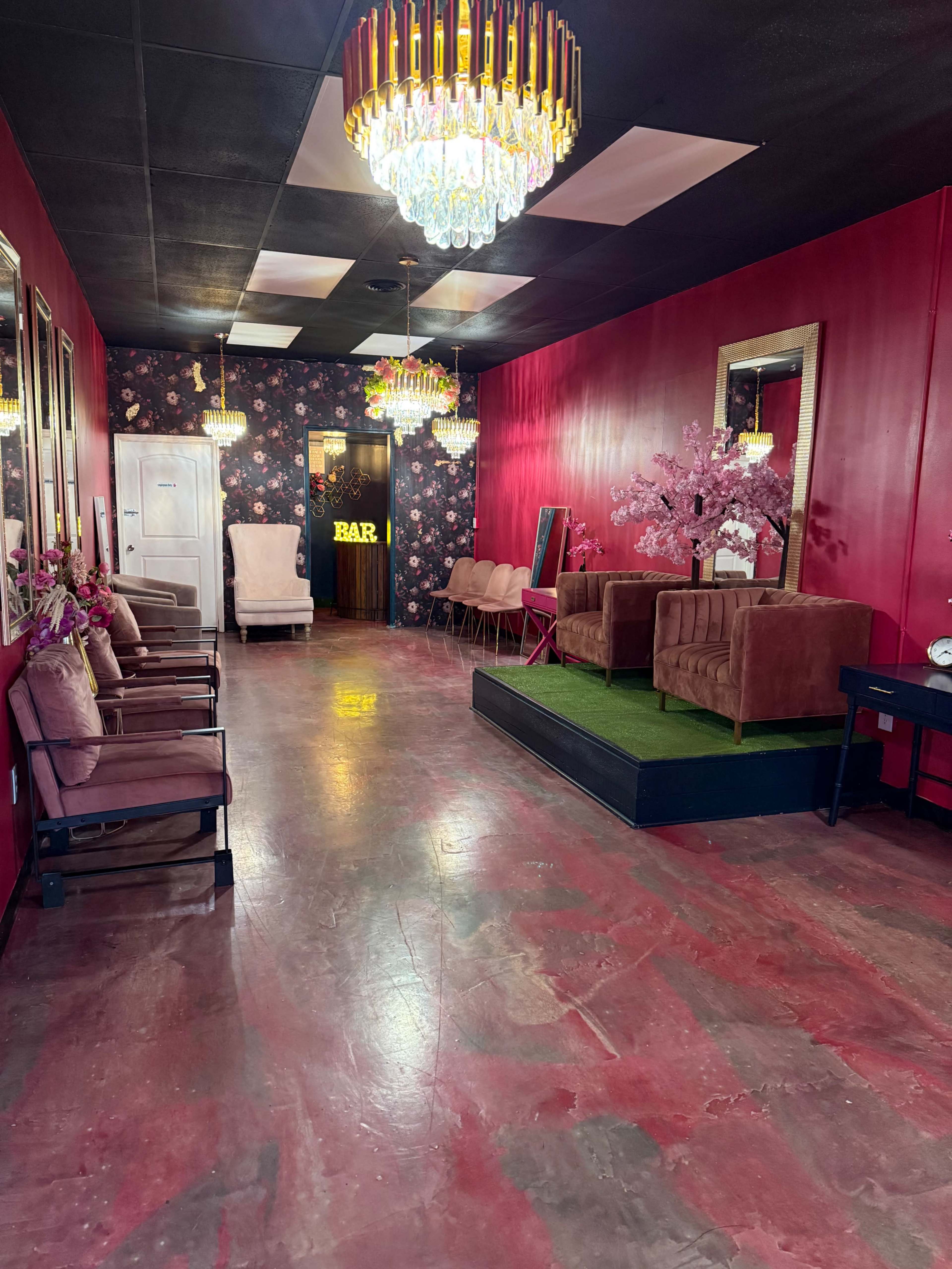 The image shows a vibrant interior space with red walls, decorative chandeliers, plush seating, and a floral theme.