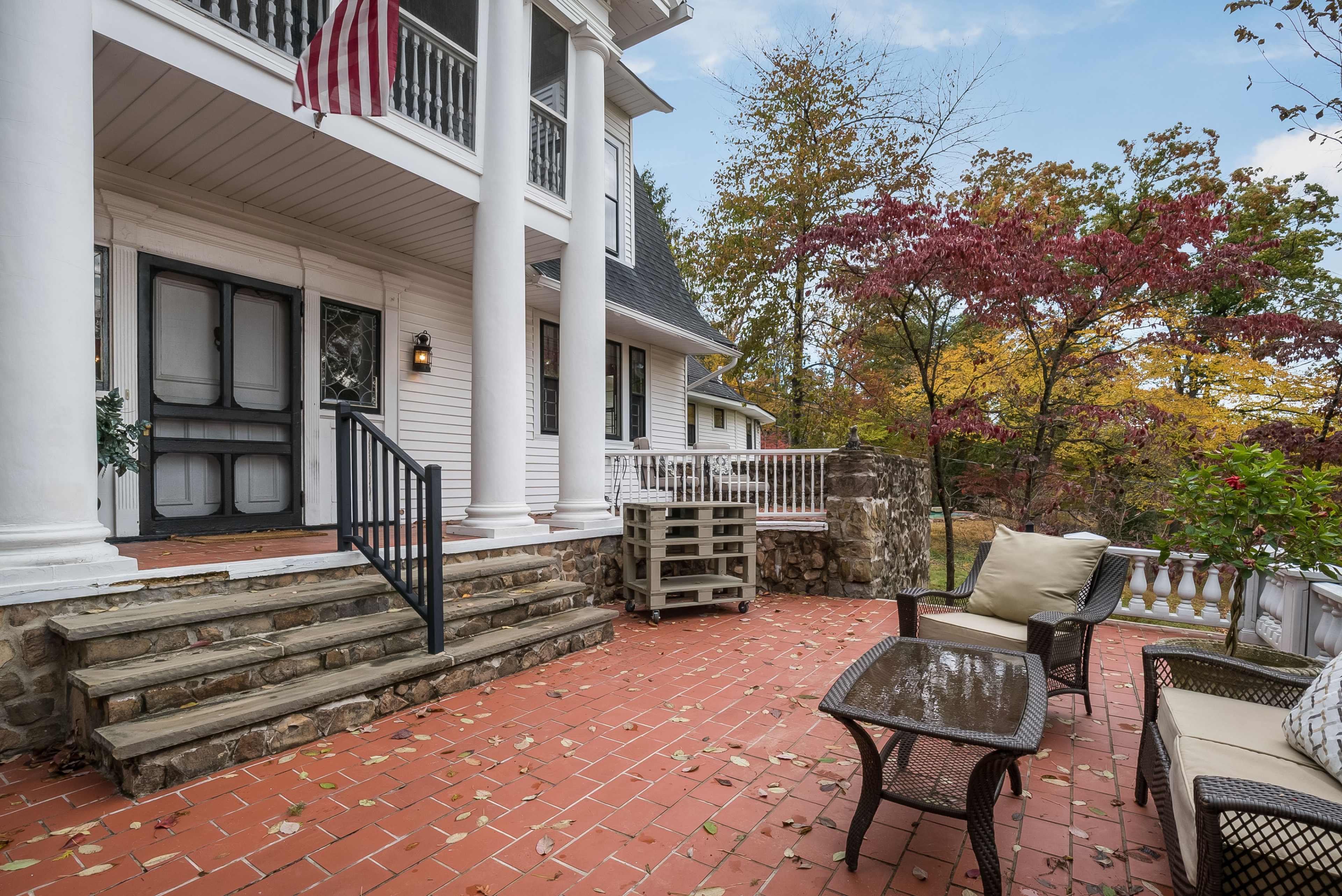 A brick patio with furniture in front of a white house surrounded by trees displaying autumn foliage.