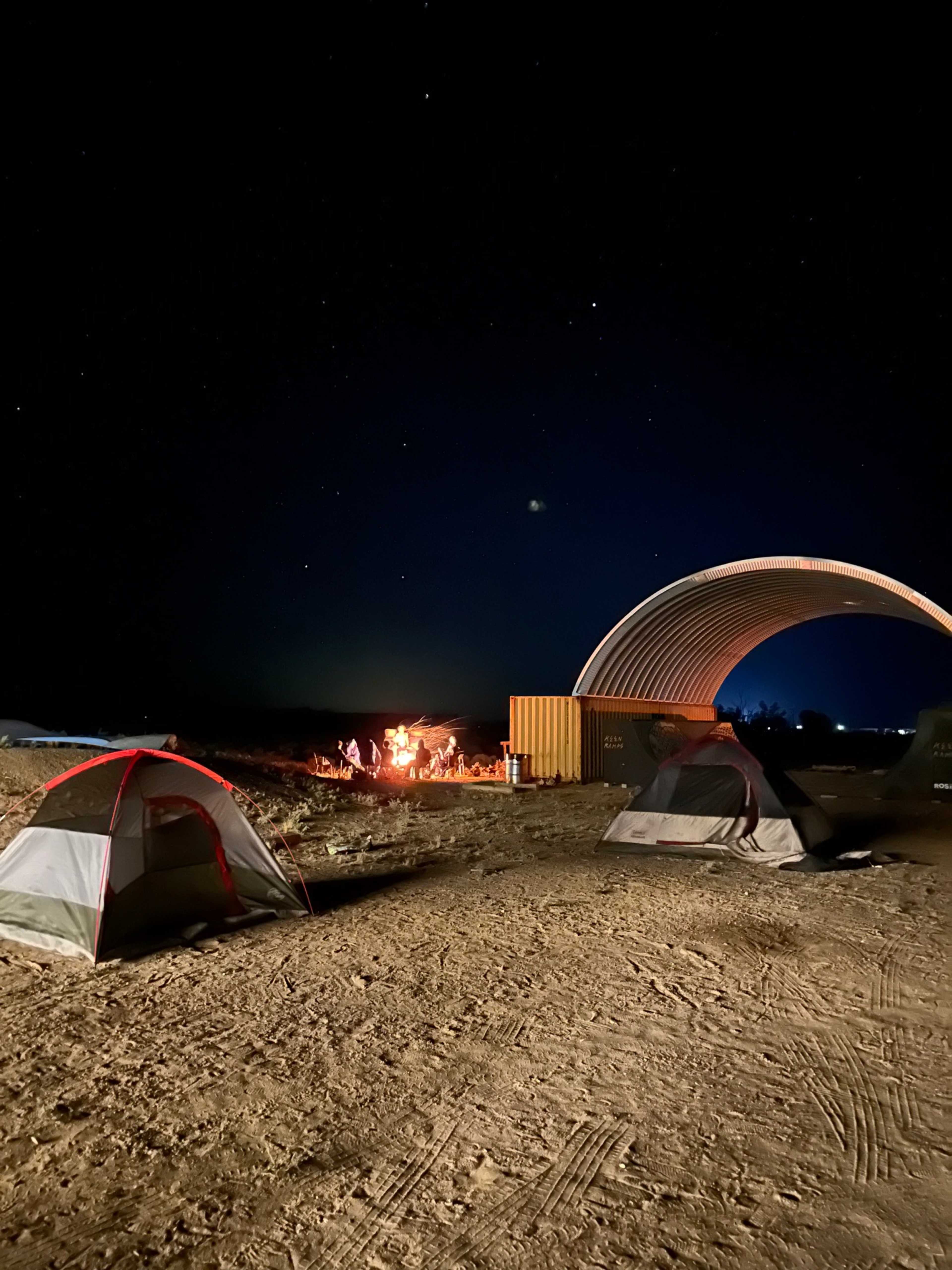 A campsite at night featuring several tents, a campfire with people gathered around it, and a structure in the background under a starry sky.