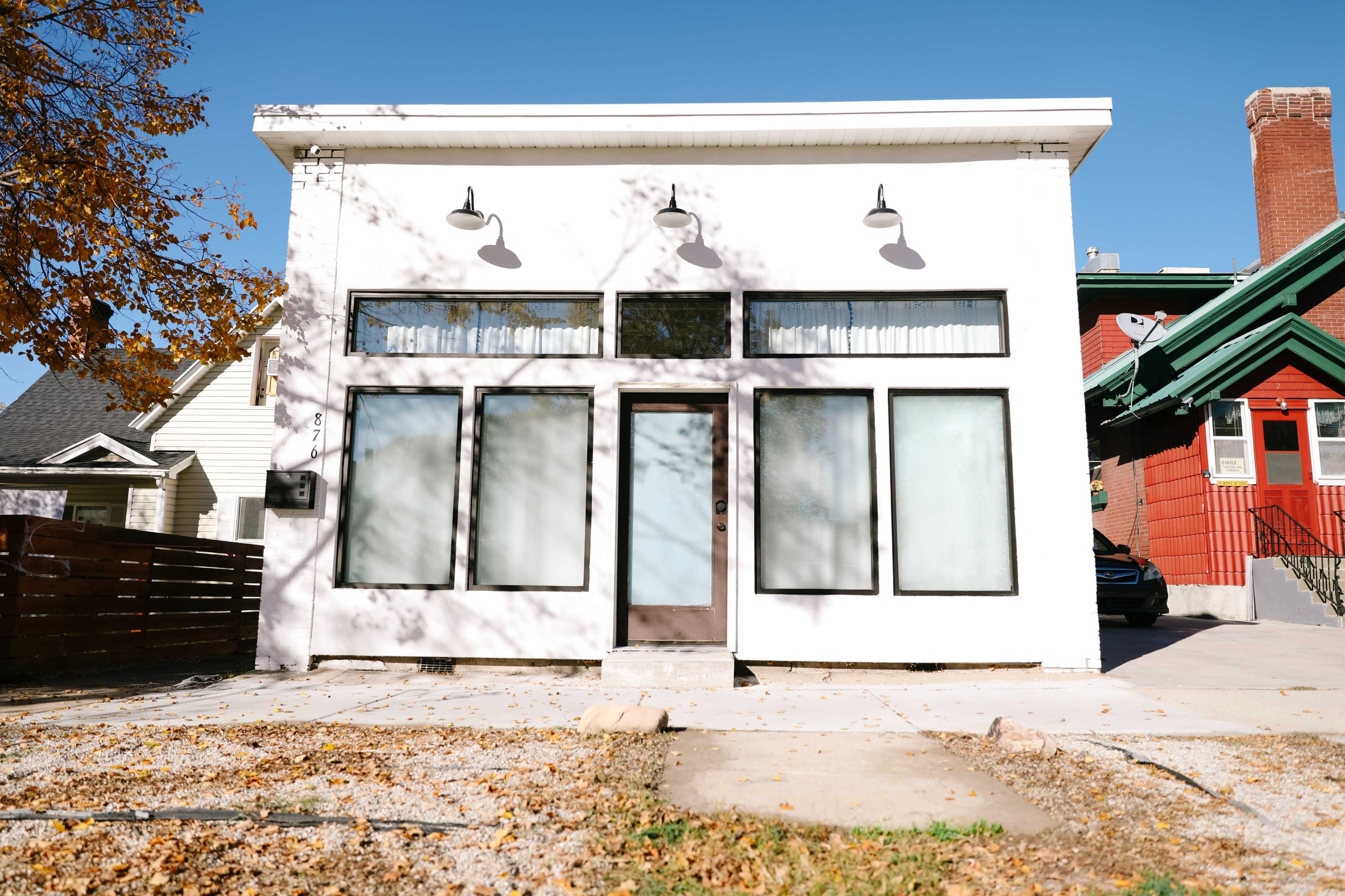 A modern white building with large windows and a central entrance is set against a clear blue sky, with fallen leaves scattered in the foreground.