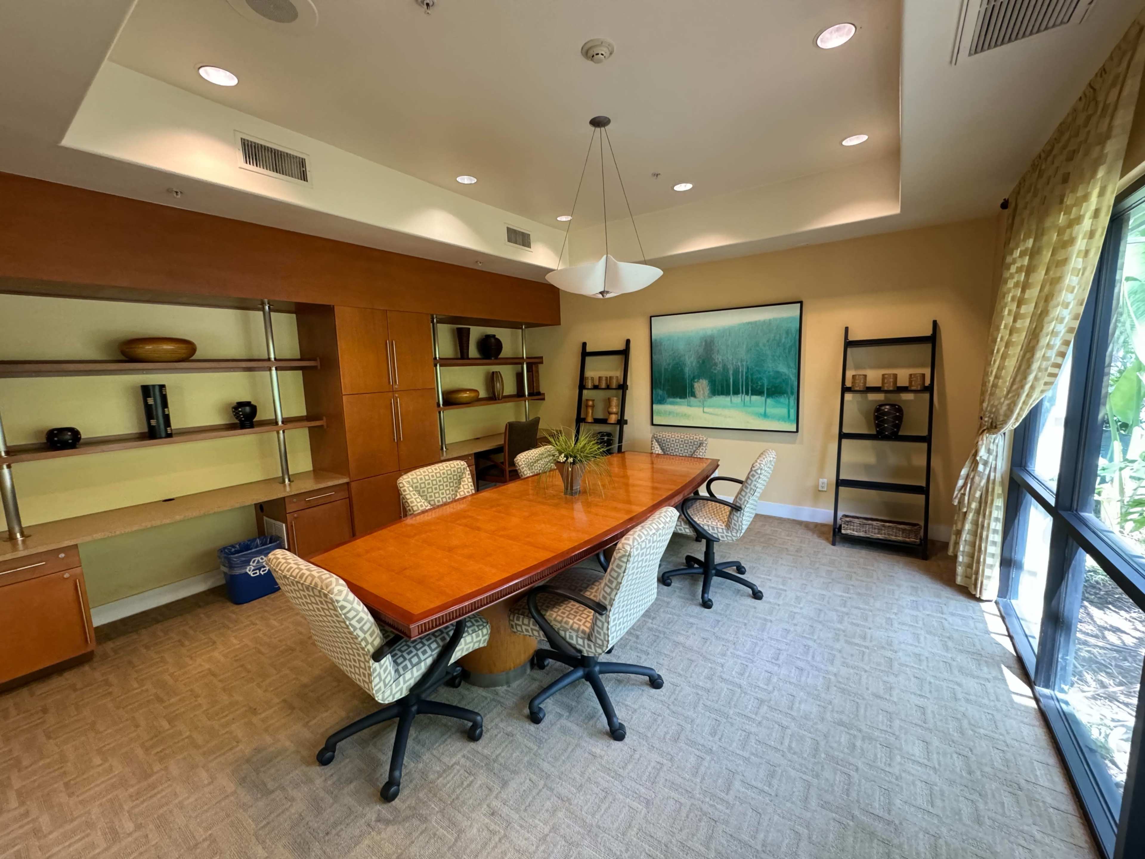 A conference room features a long wooden table surrounded by six chairs, with shelves displaying decorative items and a large artwork on the wall.