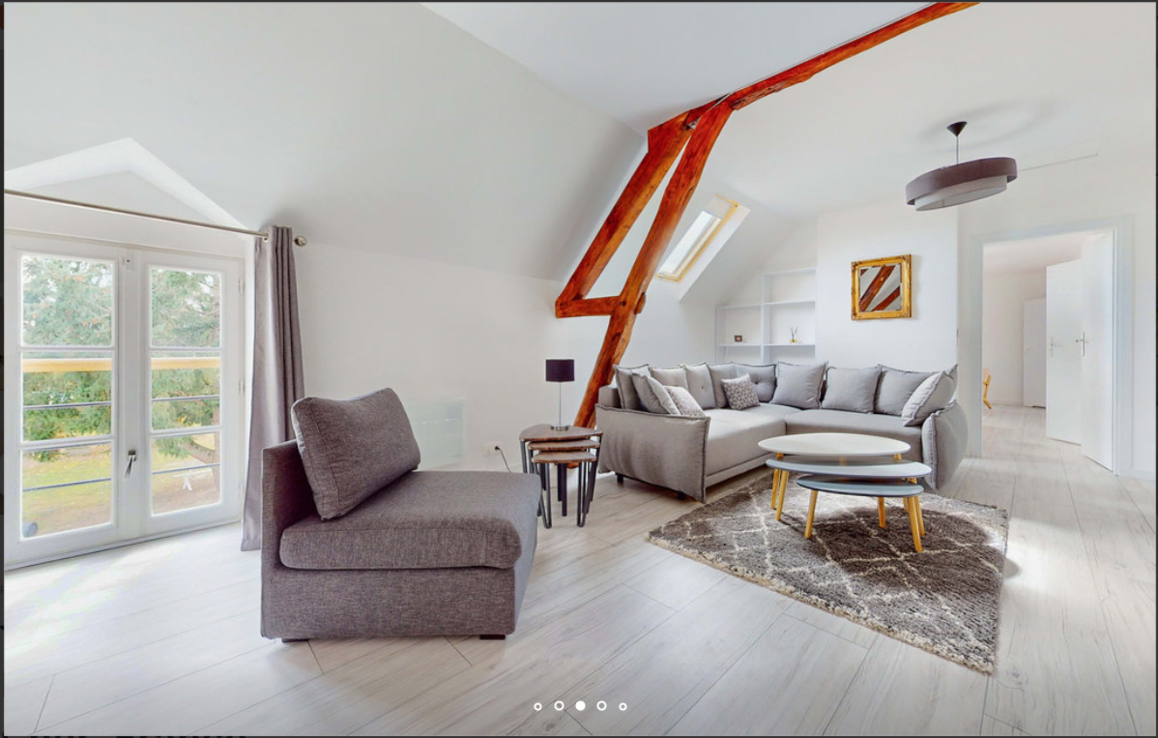 A living room features a gray sofa and armchair, a round coffee table, and large windows with a view of greenery outside.