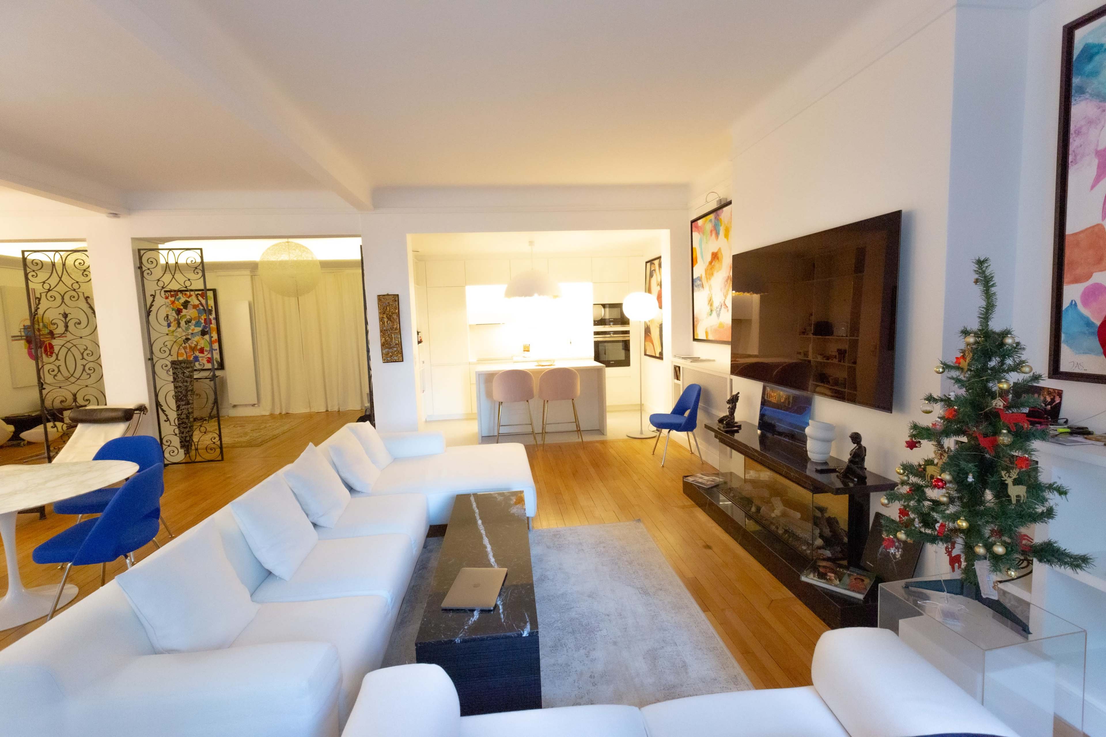 The image shows a modern living room with a white sectional sofa, a glass coffee table, a small Christmas tree, and an open kitchen in the background.
