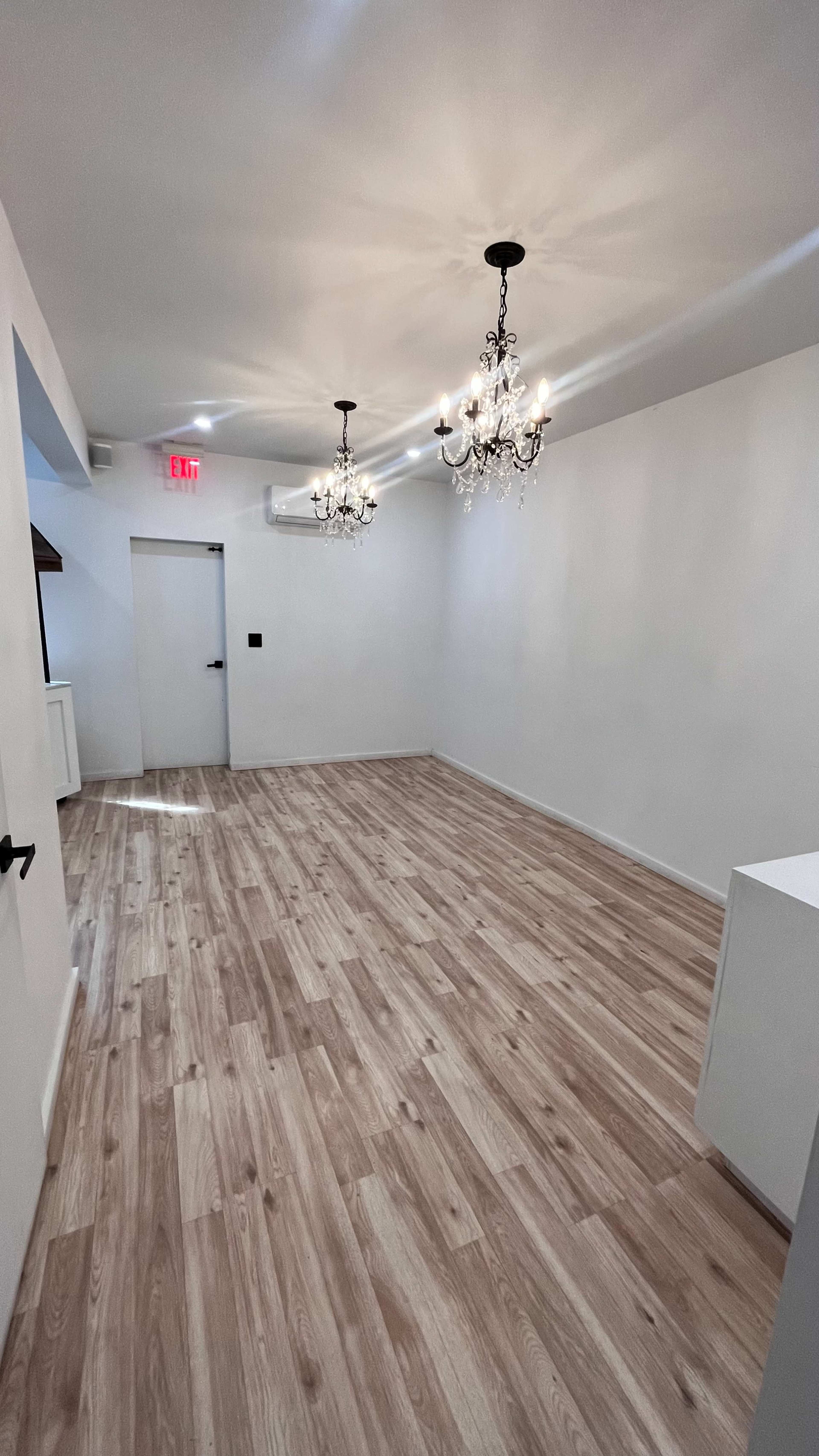 A spacious room with light wooden flooring, two black-and-gold chandeliers hanging from the ceiling, and a single door leading to another area.