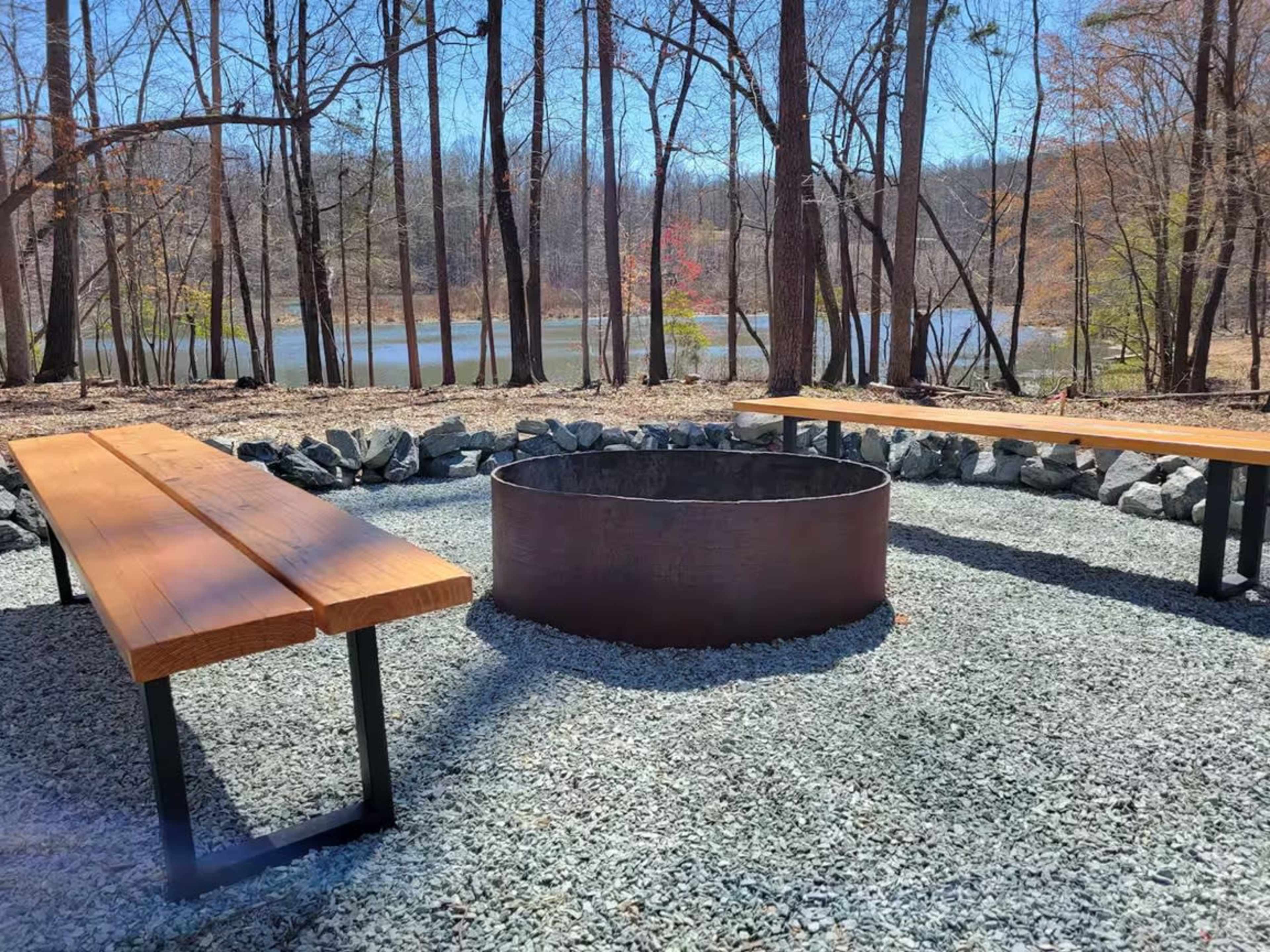 The image shows a circular fire pit surrounded by stone, with two wooden benches on a gravel surface, set against a backdrop of bare trees and a lake.