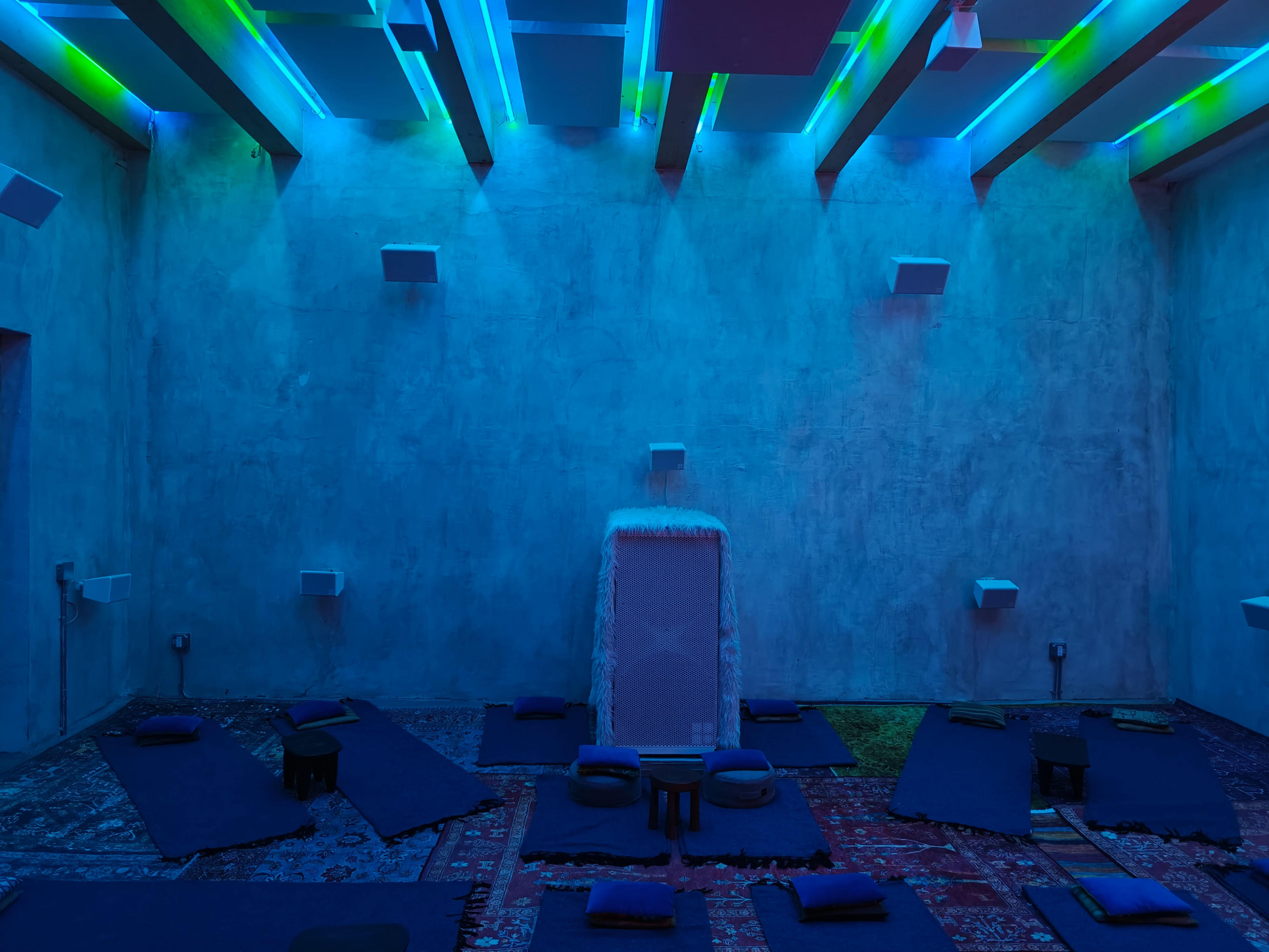 A dimly lit room features blue lighting, with yoga mats and cushions arranged on the floor against a textured concrete wall.