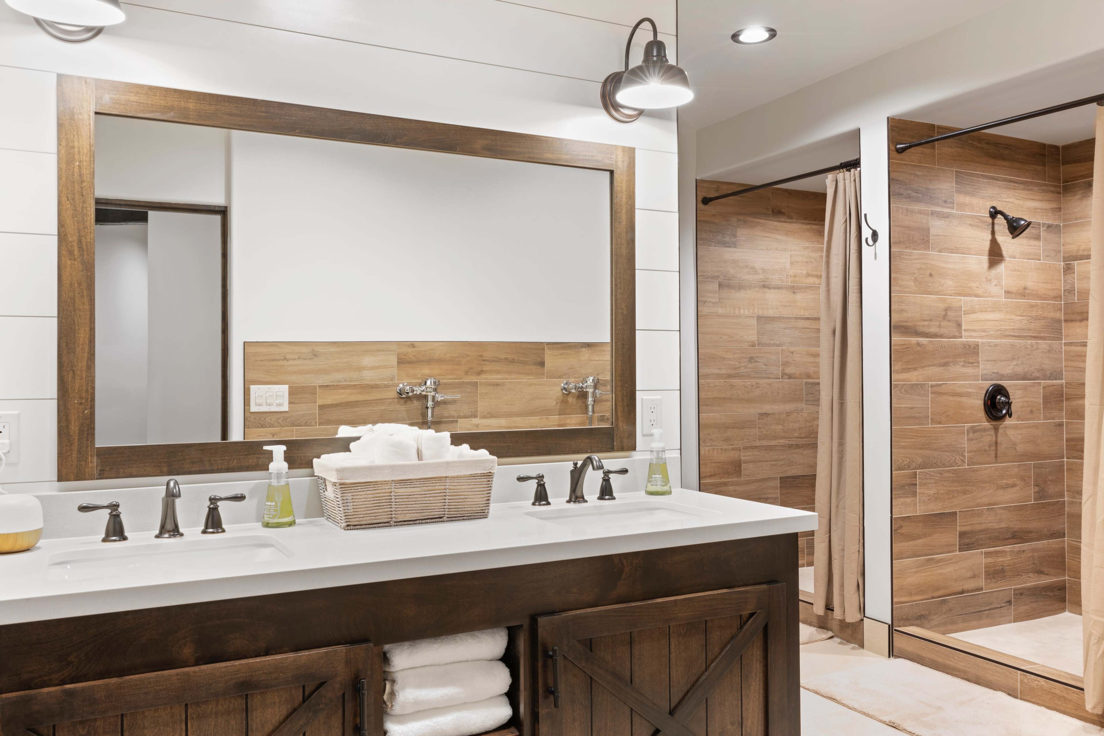 The image shows a modern bathroom with a double sink vanity featuring a large wooden mirror and a walk-in shower with wood-paneled walls.